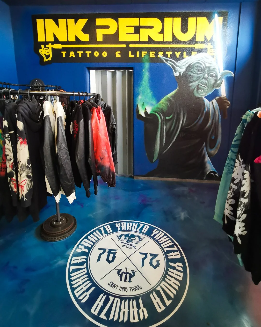 Malerie eines Yoda-ähnlichen Wesens mit einem Lichtschwert vor einer blauen Wand. Kleidung auf Kleiderständern im Vordergrund. Text auf dem Boden und an der Wand, Werbung für Ink Perium-Tattoo- und Lifestyle-Studio. Professionelle Graffitigestaltung 