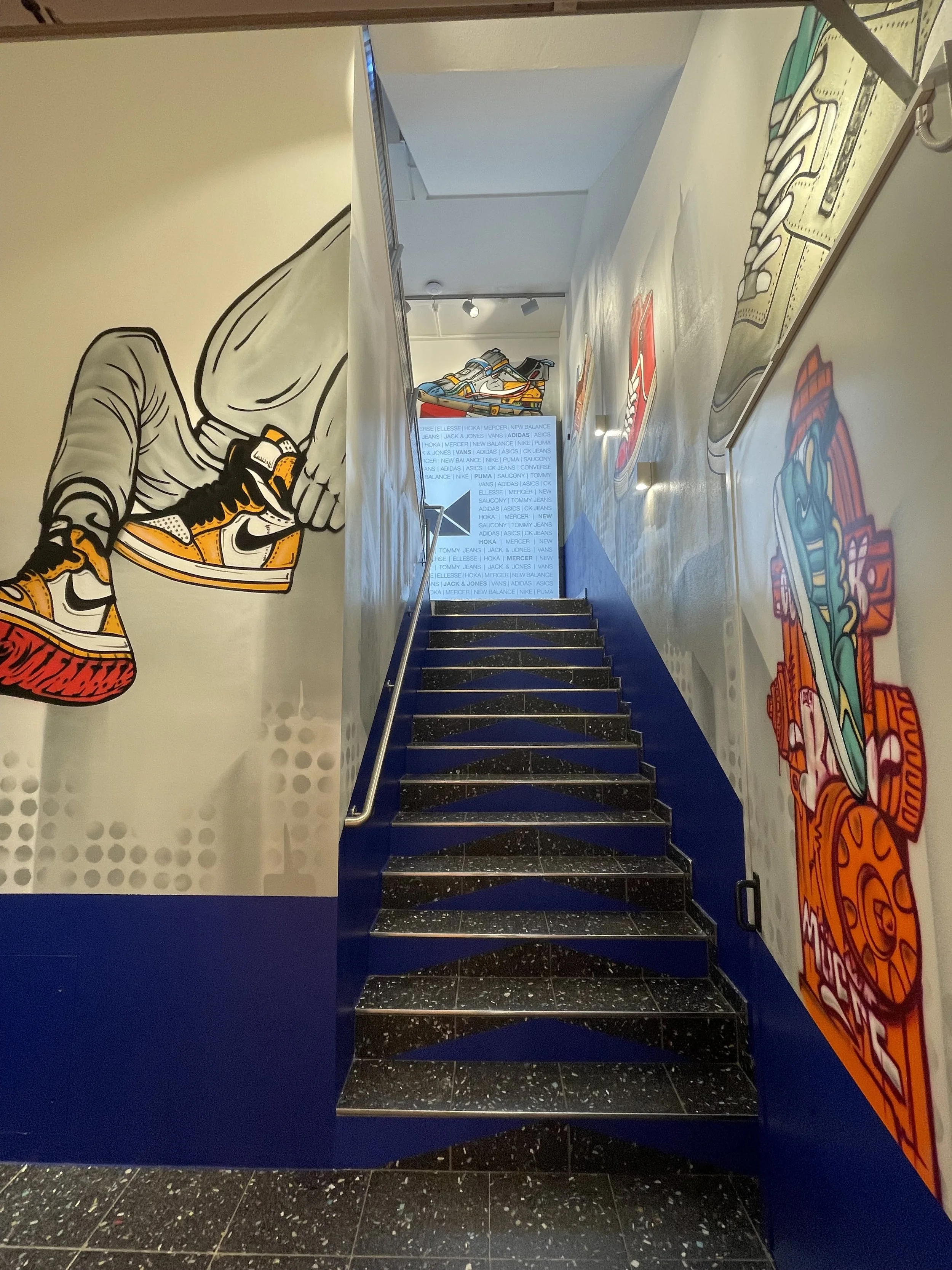 Treppen in einem inneren Bereich mit Wandbildern von Sportschuhen und einer Wand mit einem Digitalbildschirm im Hintergrund.