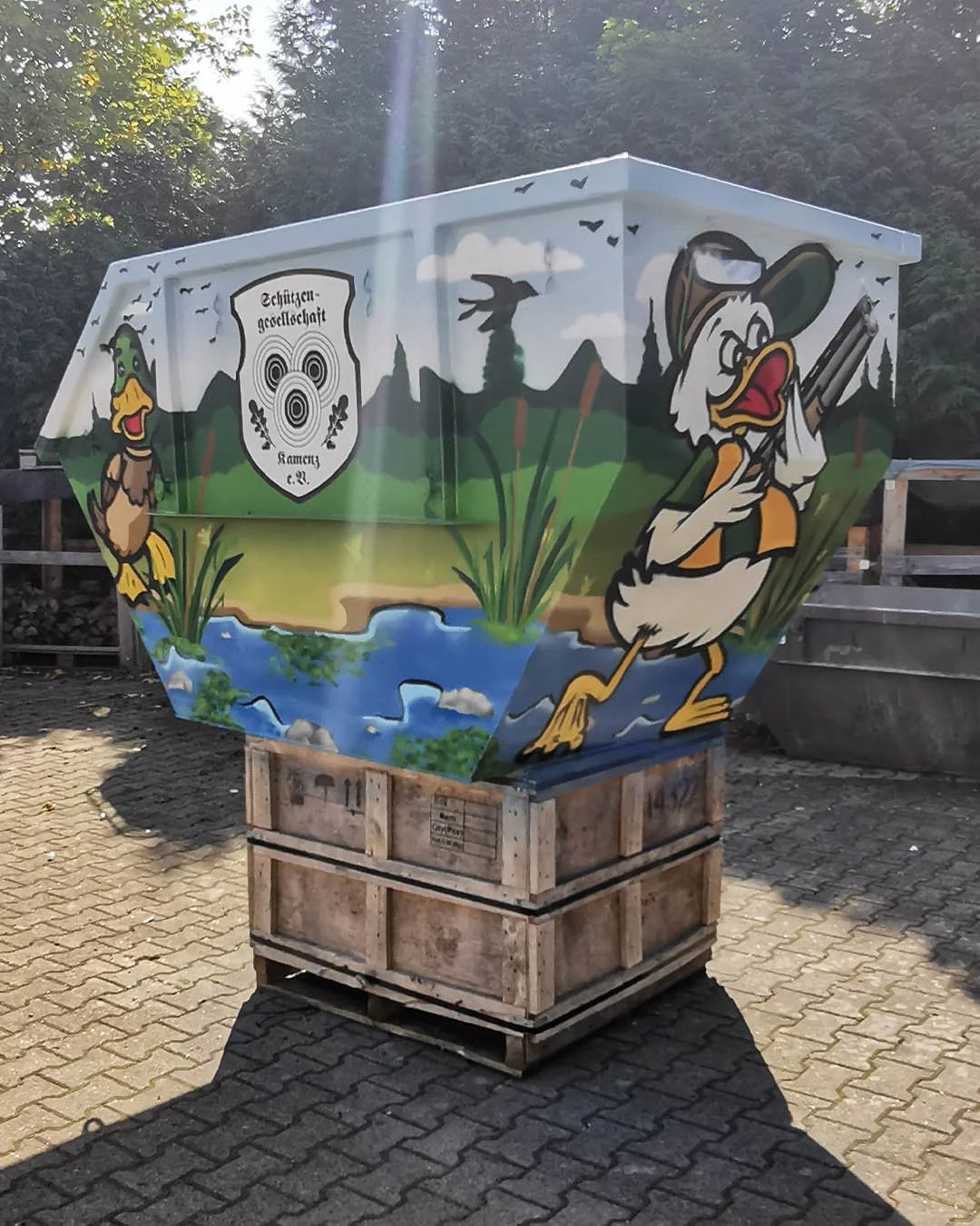 Ein bemalter Truhenwagen mit einem Wasser- und Naturlandschaft und Comic-Charakteren, darunter eine Ente mit Piratenhut und eine andere Ente mit Schnabel und Flossen, die eine Angel hält, sowie ein Streifenwappen mit der Aufschrift 'Schützen-Gesellsc