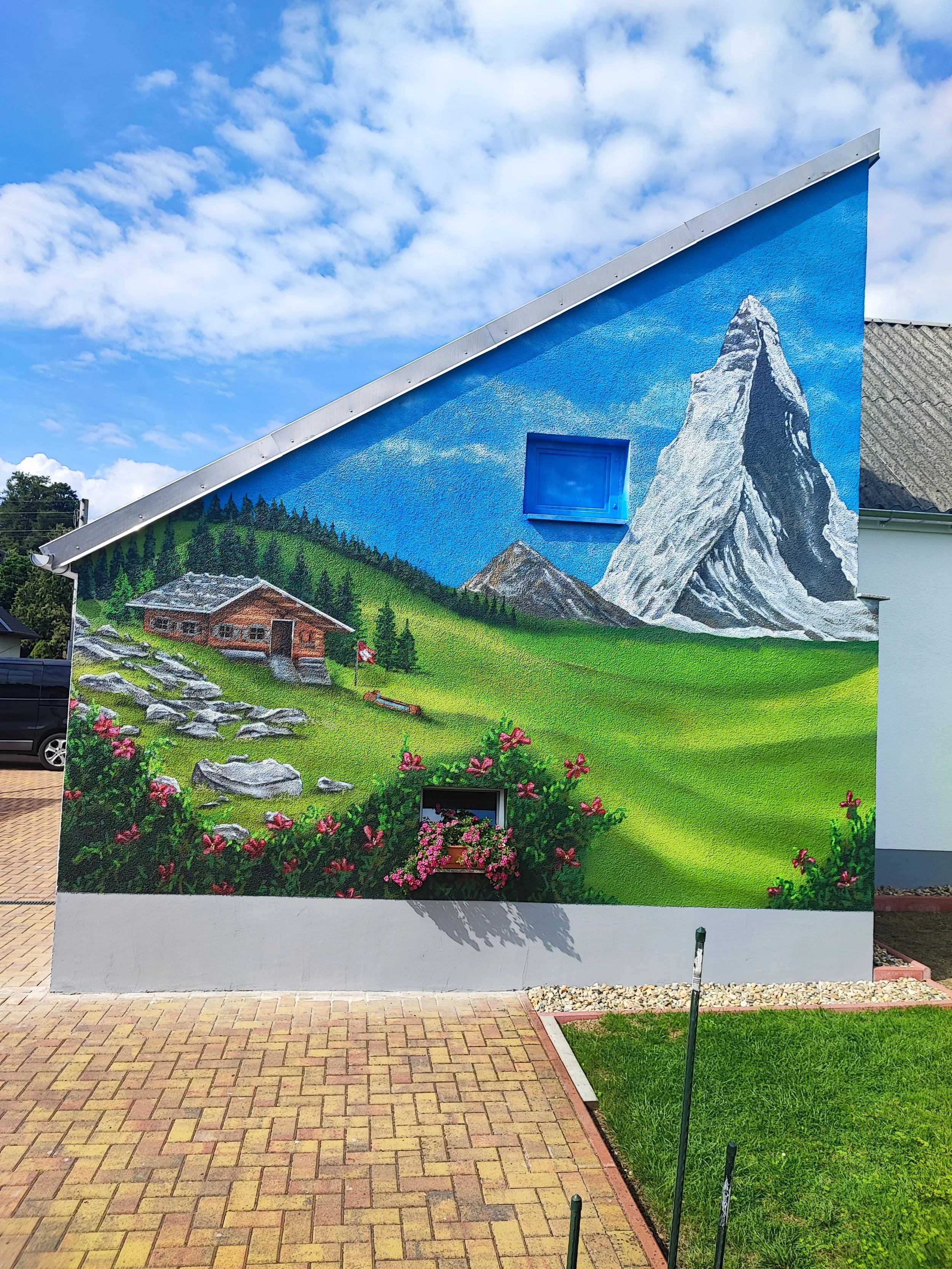 Wandmalerei auf einem Gebäude, die eine Alpenlandschaft mit einem großen Berg, einer kleinen Hütte, grünen Wiesen und Blumen zeigt, mit blauer Himmel und Wolken im Hintergrund.
