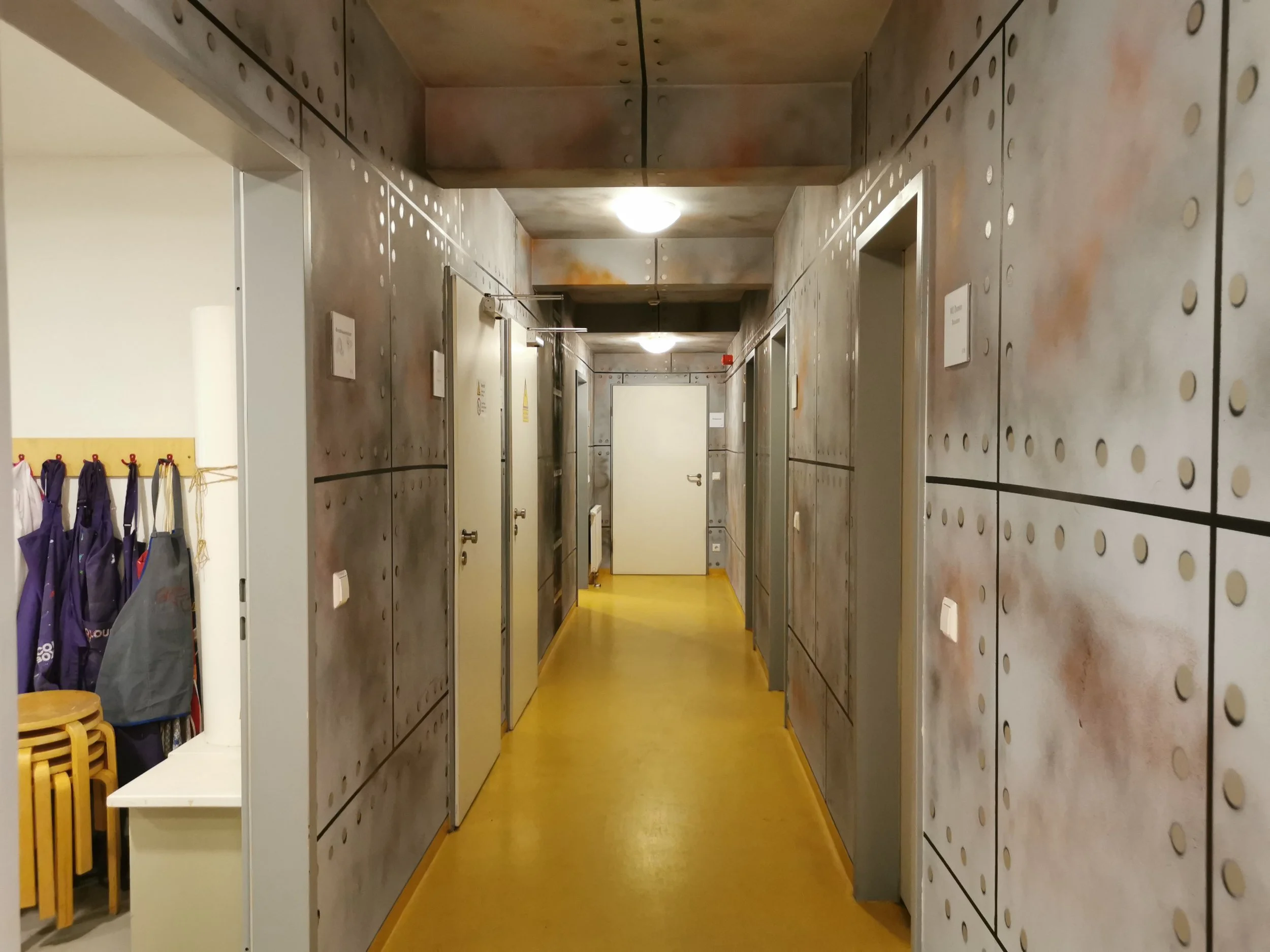 Innenansicht einer Beton-Gang mit mehreren Türen und einem gelben Boden. An der linken Seite sind Garderoben mit eingepackten Regenschirmen. Die Wände sind mit Metallplatten verkleidet.