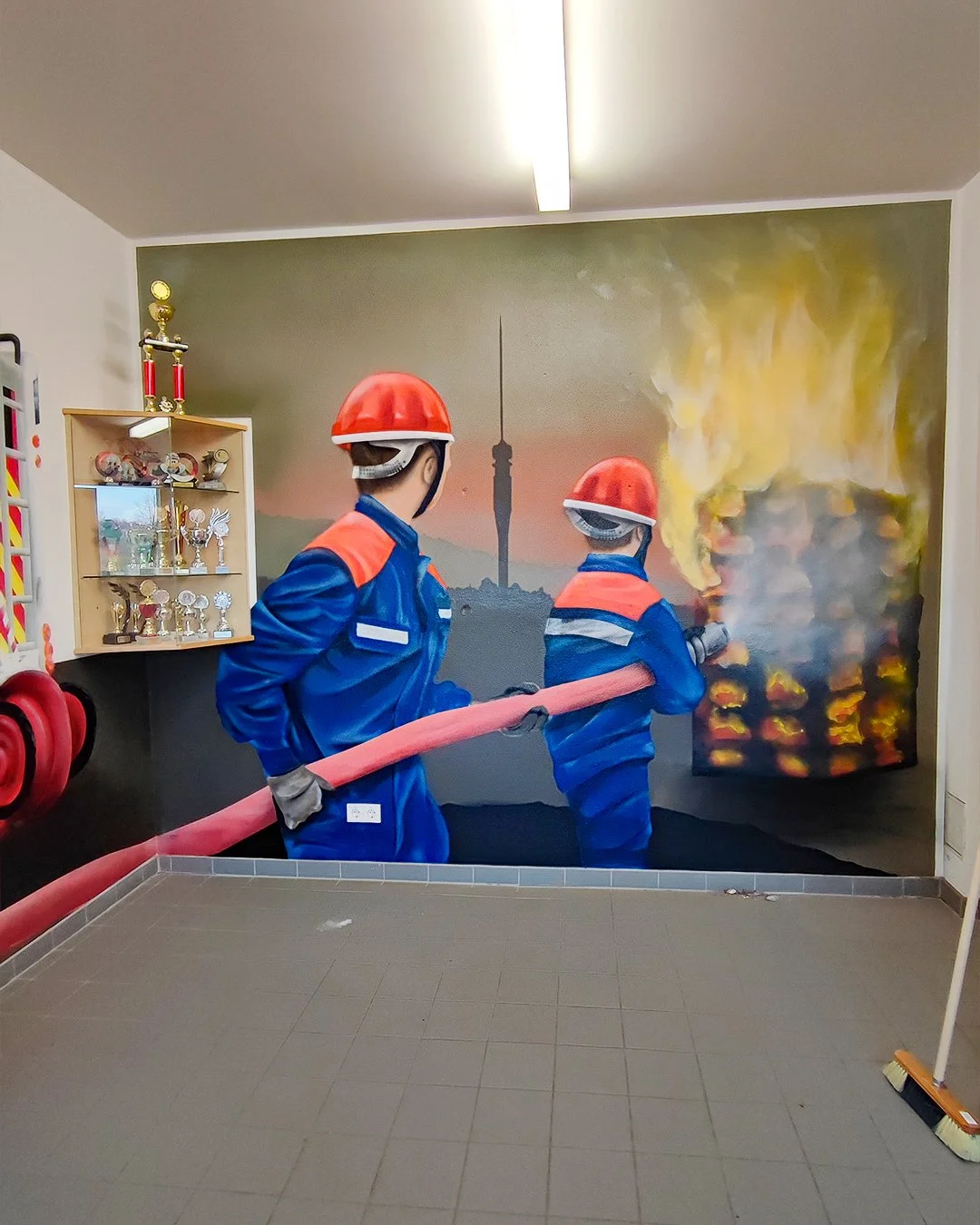 Wandbemaltes Bild von zwei Feuerwehrleuten, die einen Brand mit einem Schlauch löschen, mit einem Stadtbild und einem Fernsehturm im Hintergrund.