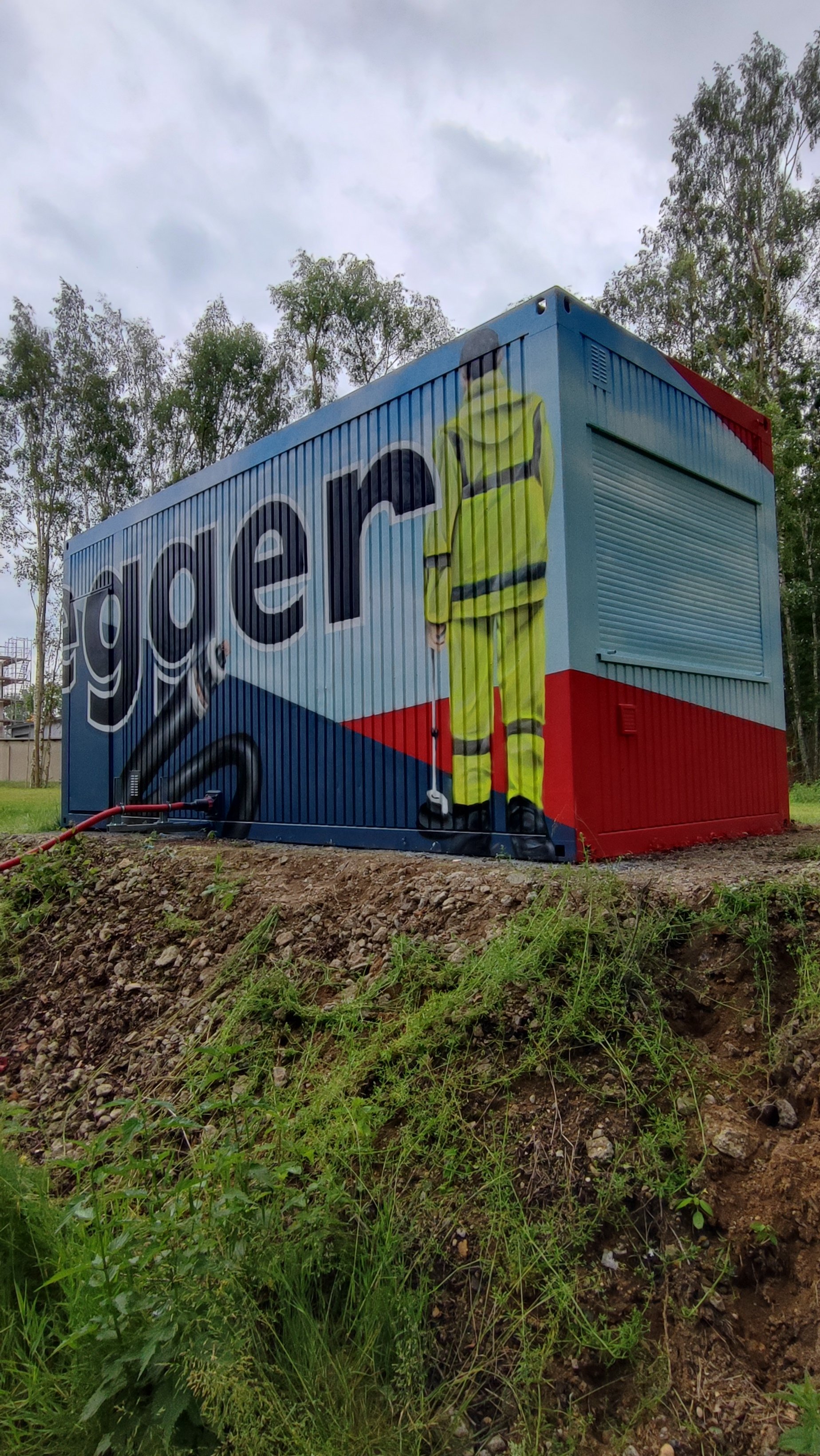 Graffiti-Kunst auf einem kleinen blauen und roten Container mit einem Bauarbeiter in Leuchtgelb. Im Hintergrund sind Bäume und ein bewölkter Himmel.