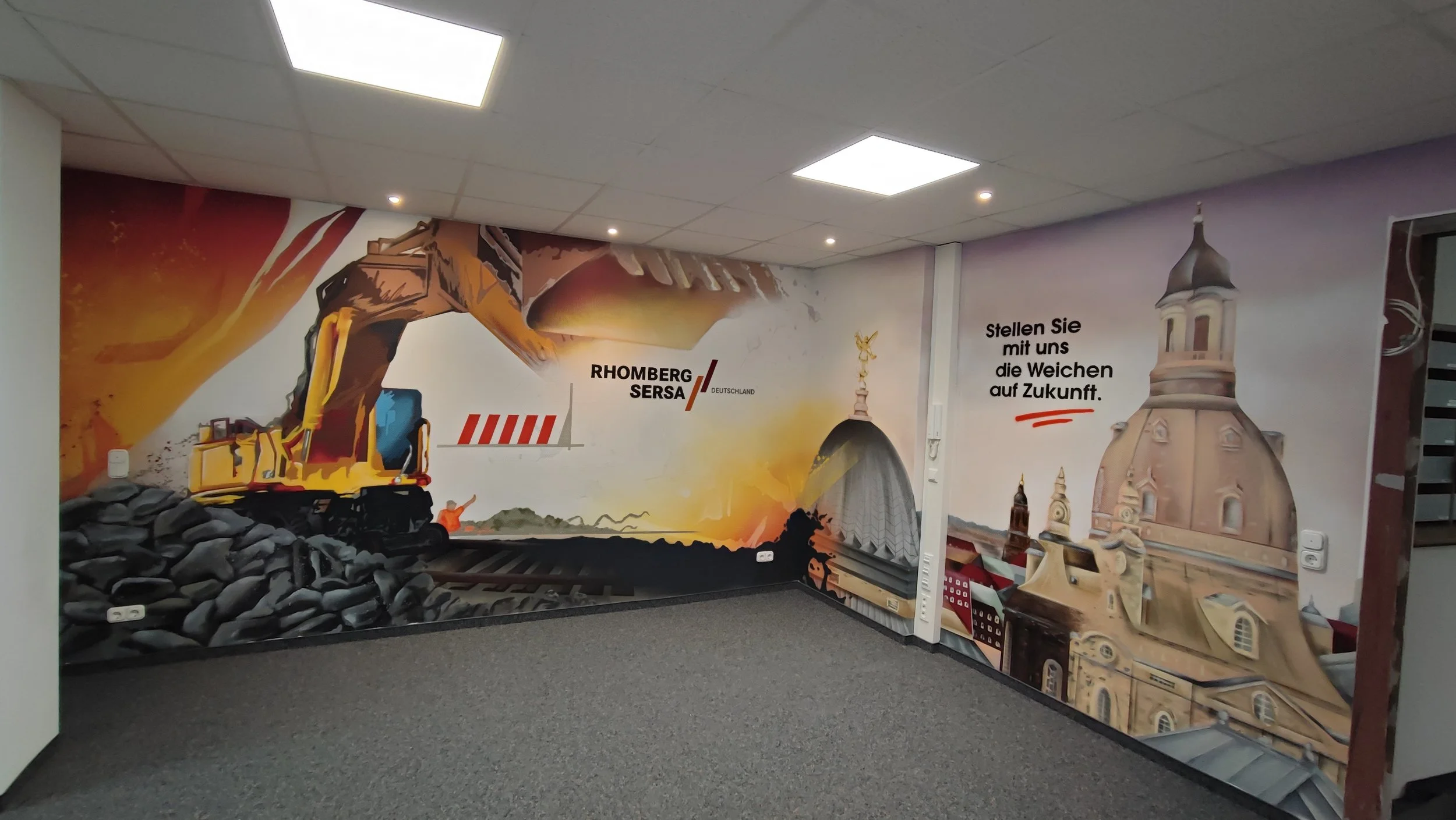 Wandmalerei in einem Raum, die eine Baustelle mit einem Bagger, den Namen 'Rhomberg Sersa Deutschland', und historische Gebäude sowie eine Kirche zeigt. Text auf der Wand lautet: 'Stellen Sie mit uns die Weichen auf Zukunft'. Professionelle Graffitig