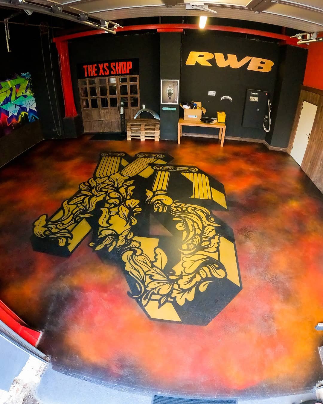 Ein leerer Raum mit graffiti-artigem Boden, einer Wand mit einem gelben „RWB“-Logo und „THE XS SHOP“-Schriftzug, Holzpaletten, einem kleinen Tisch, einem Schrank und einer Graffiti an der linken Wand.