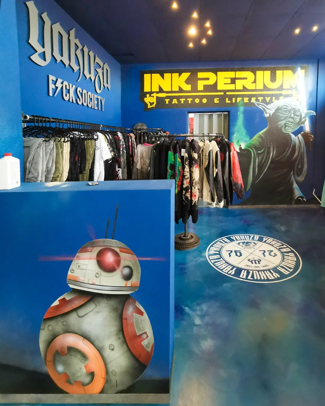Innenraum eines Ladens mit Kleidung auf Kleiderstangen, eine Wand mit Graffiti und einem großen Yoda von Star Wars, sowie einen blauen Wandbereich mit dem Schriftzug 'INK PERIUM Tattoo & Lifestyle' und einem weiteren Schriftzug 'Yakuza F CK Society'.