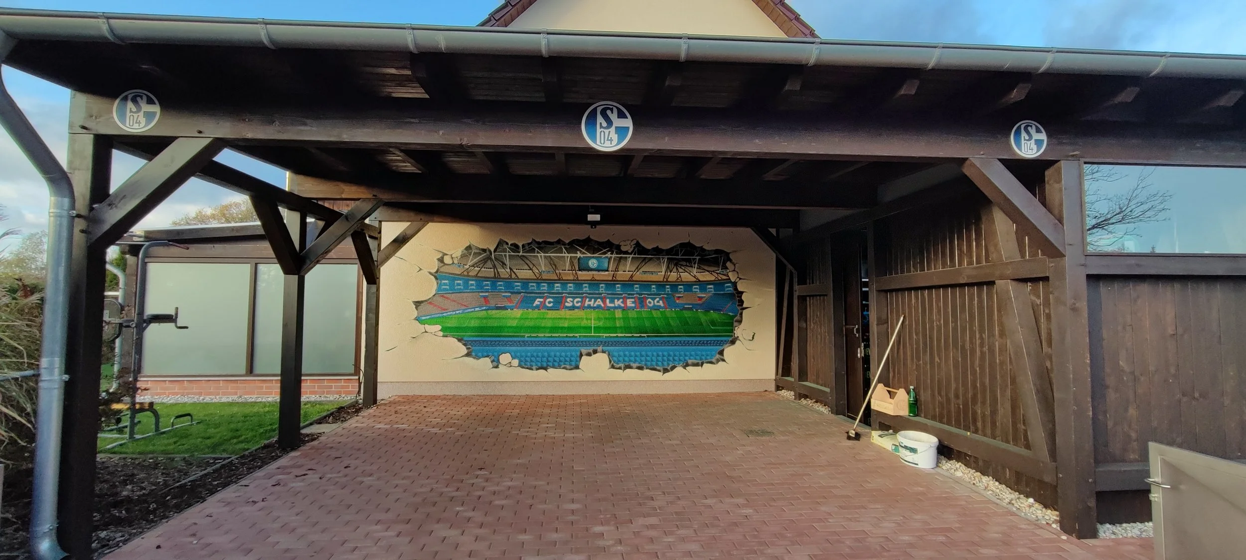 Eine Wanddekor mit dem Logo des FC Schalke 04, das aussieht, als ob eine Wand durchbrochen wurde, um das Stadion im Inneren zu zeigen.