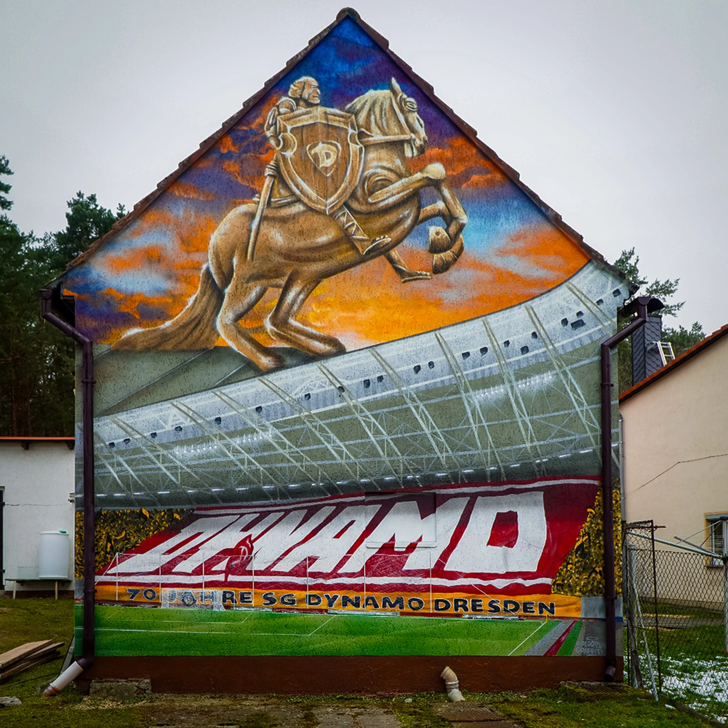 Ein Wandbild eines Ritters auf einem galoppierenden Pferd mit einem Schild, im Hintergrund ein Himmel mit Orange und Blau, darunter ein Banner mit dem Begriff 'Dynamo Dresden' und weiteren Texten.