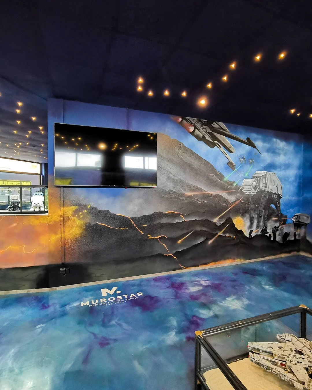 Mural mit Science-Fiction-Thema, zeigt Raumschiff, Kampfroboter und Zerstörung auf einer Landschaft mit Lava. Firmenname 'MuroStar' unten an der Wand, Innenraum mit gläsernem Ausstellungsstück und Fernseher.