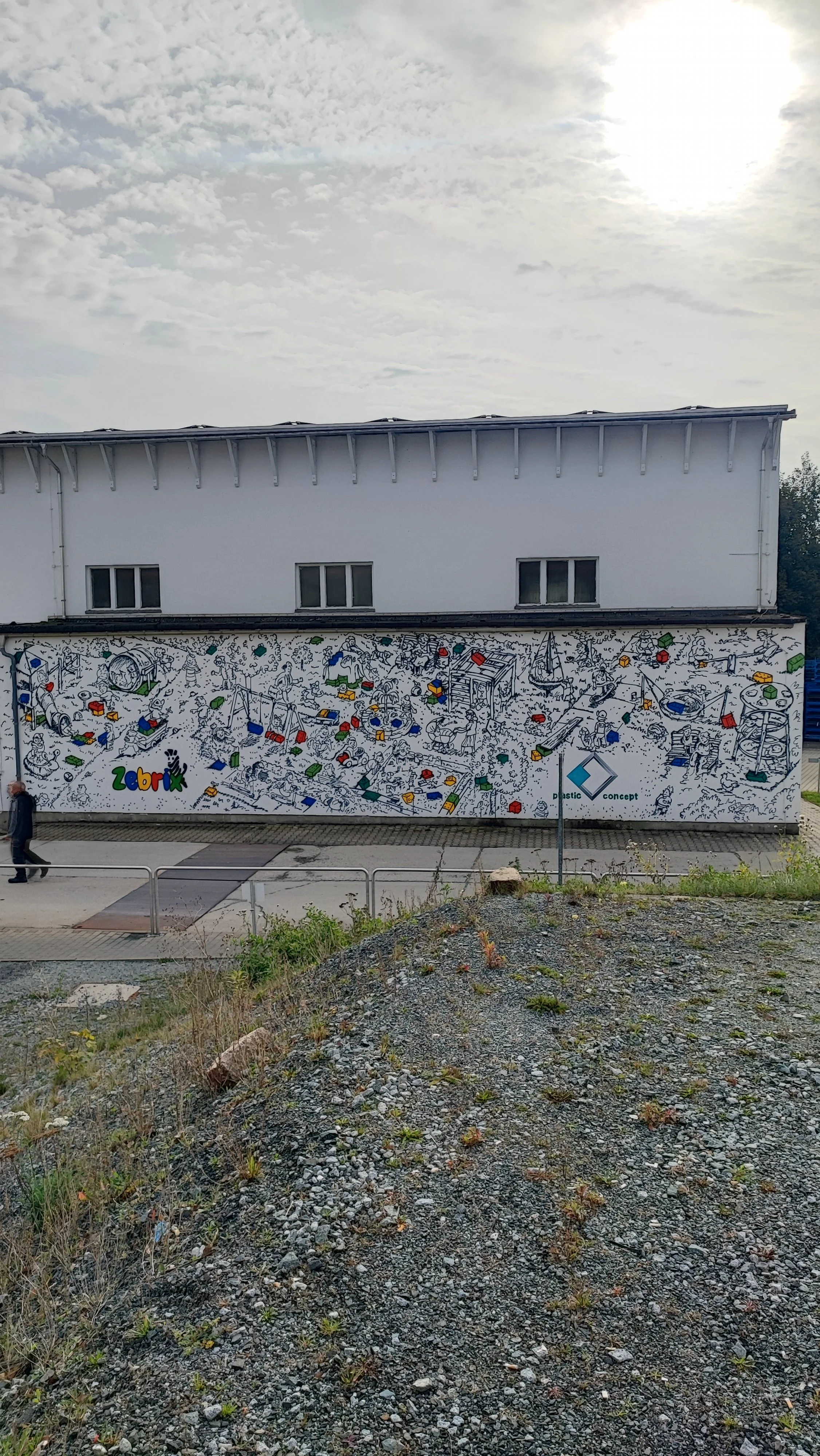 Wandgemälde in Schwarz-Weiß mit bunten Akzenten, zeigt eine fantasievolle Kinderwelt mit Spielzeug, Fahrzeugen und Kindern, darunter der Name »Zebrix«. Logo von 'plastic concept' ist sichtbar.