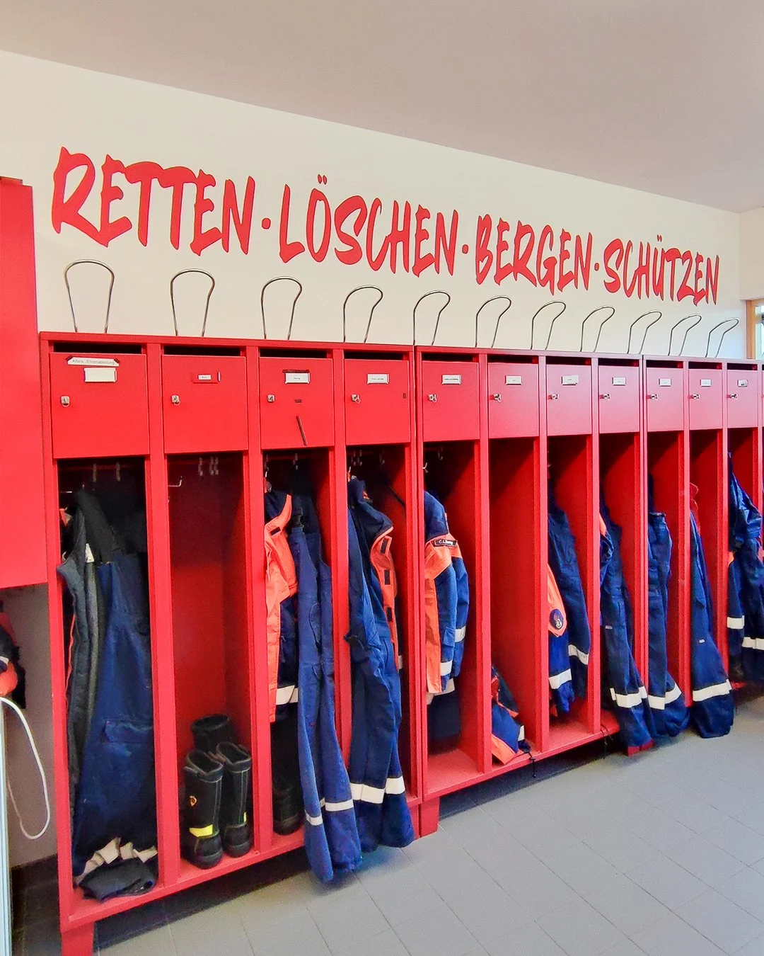 Rote Schließfächer mit Feuerwehrjacken und -stiefeln in einem Flur. Über den Schließfächern steht auf der Wand in roter Schrift: Rettung, Löschen, Bergen, Schützen.