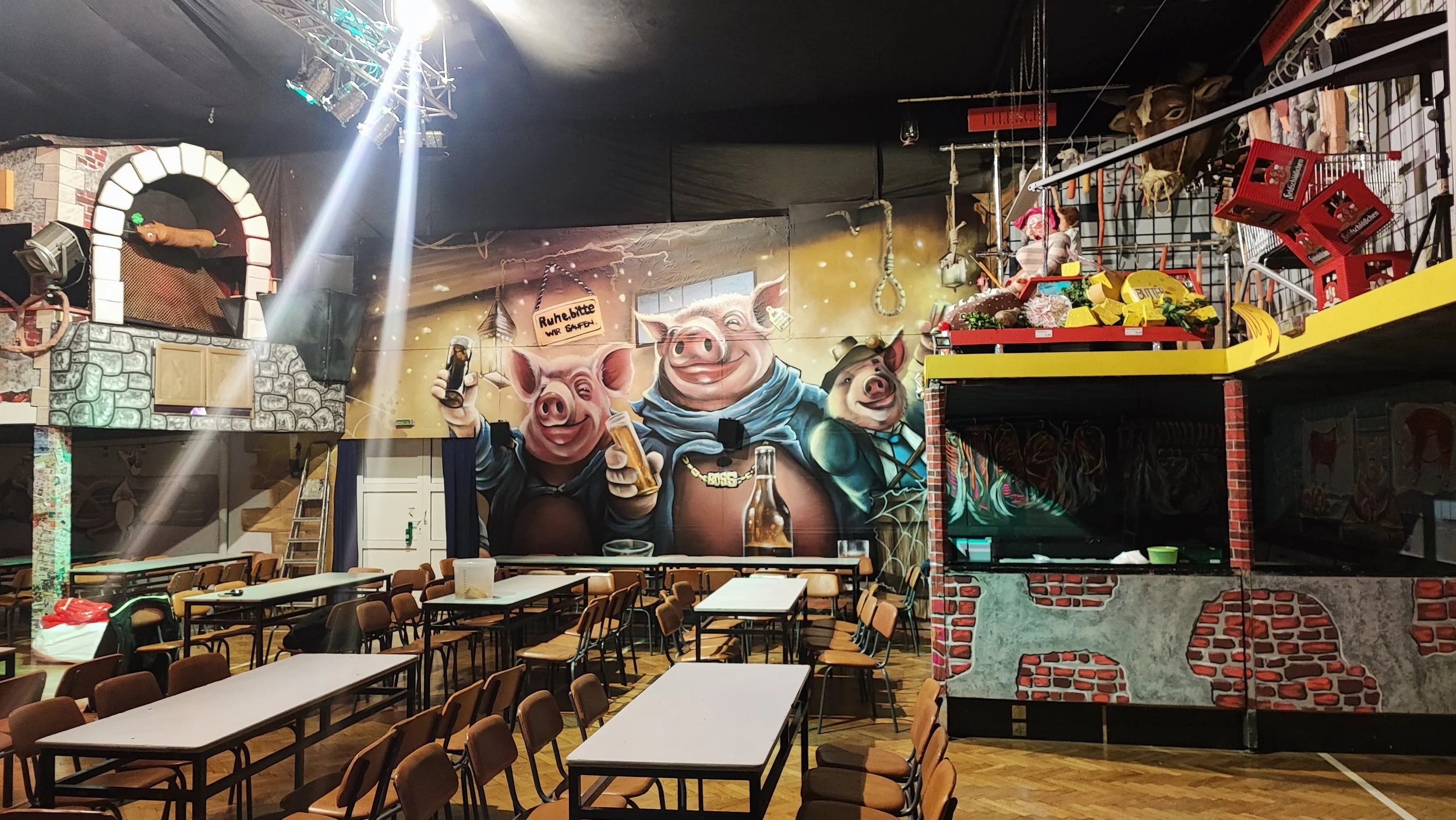 Innenraum eines Restaurants mit Tafelwand, auf der drei fröhliche Schweine geschminkt sind, die Bier trinken, und ein Spielbereich mit Rutschen und Spielsachen.