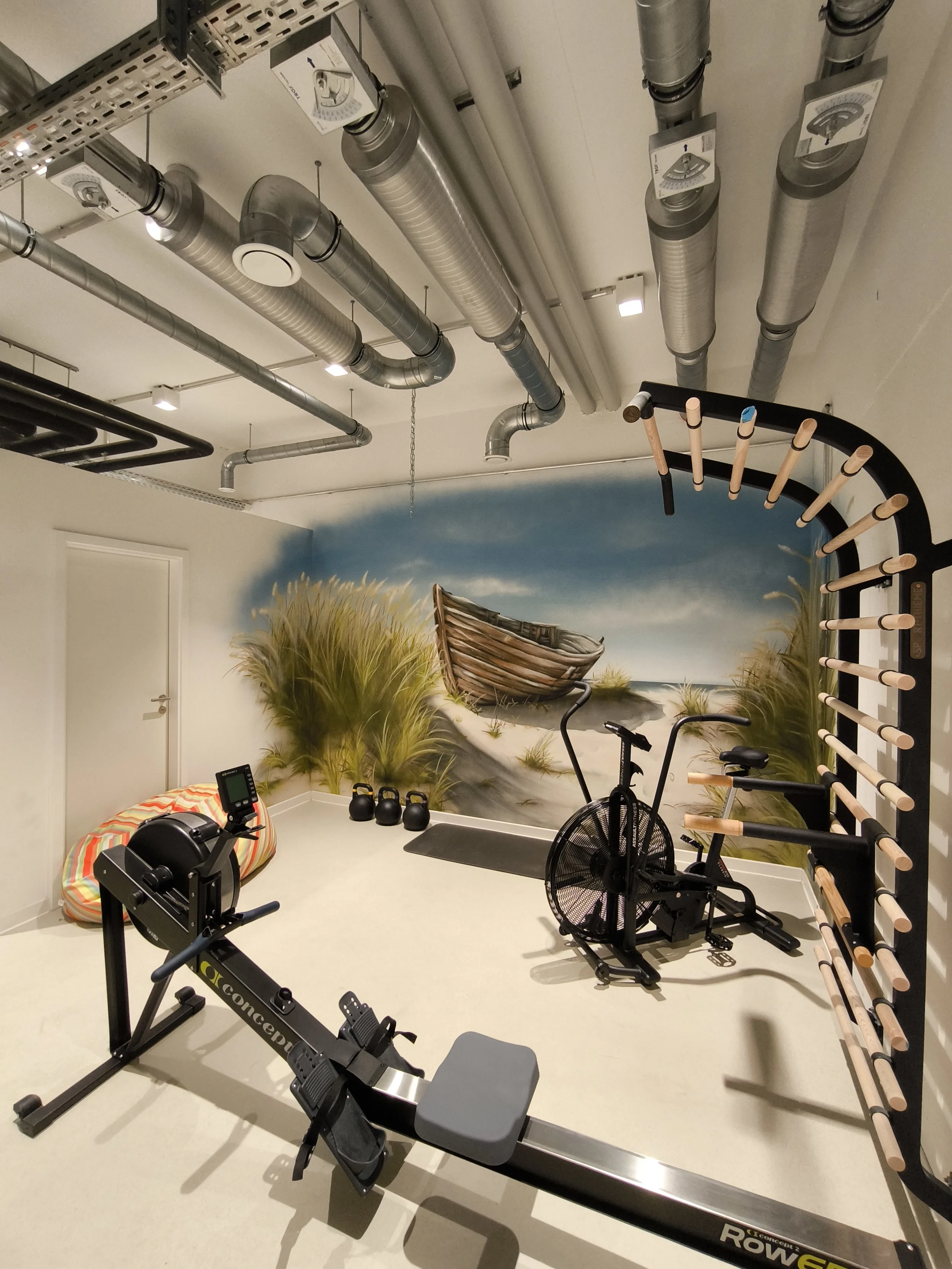 Ein Fitnessraum mit Wandmalerei eines Bootes am Strand, zwei Heimtrainer, ein Rudergerät, eine Kletterwand und verschiedene Trainingsgeräte. Die Decke hat sichtbare Lüftungsschächte.
