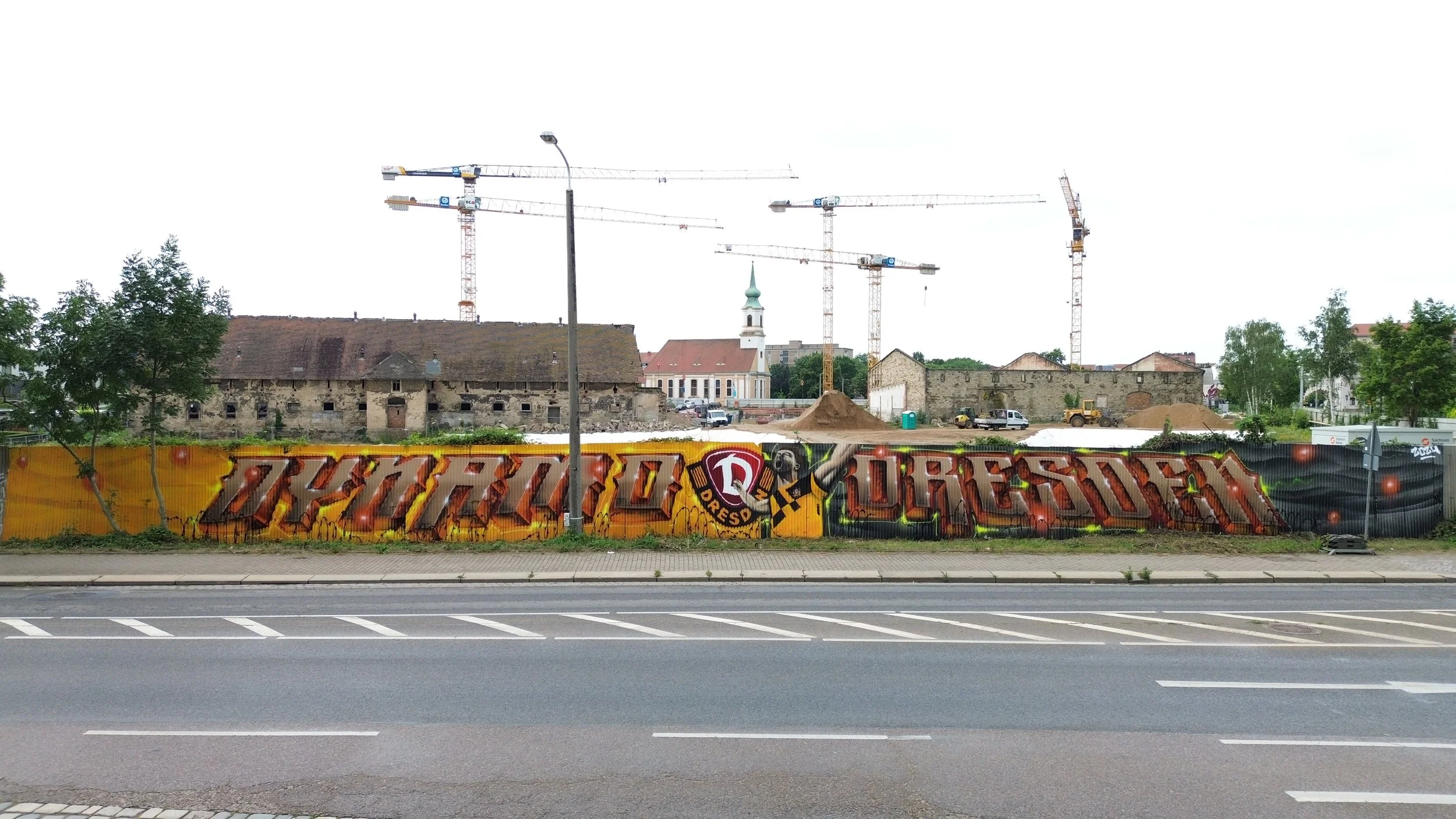 Bauzaun mit Graffiti, Bauarbeiten mit Kräne und Baufahrzeuge, alte Gebäude im Hintergrund, Kirche mit Turmuhr