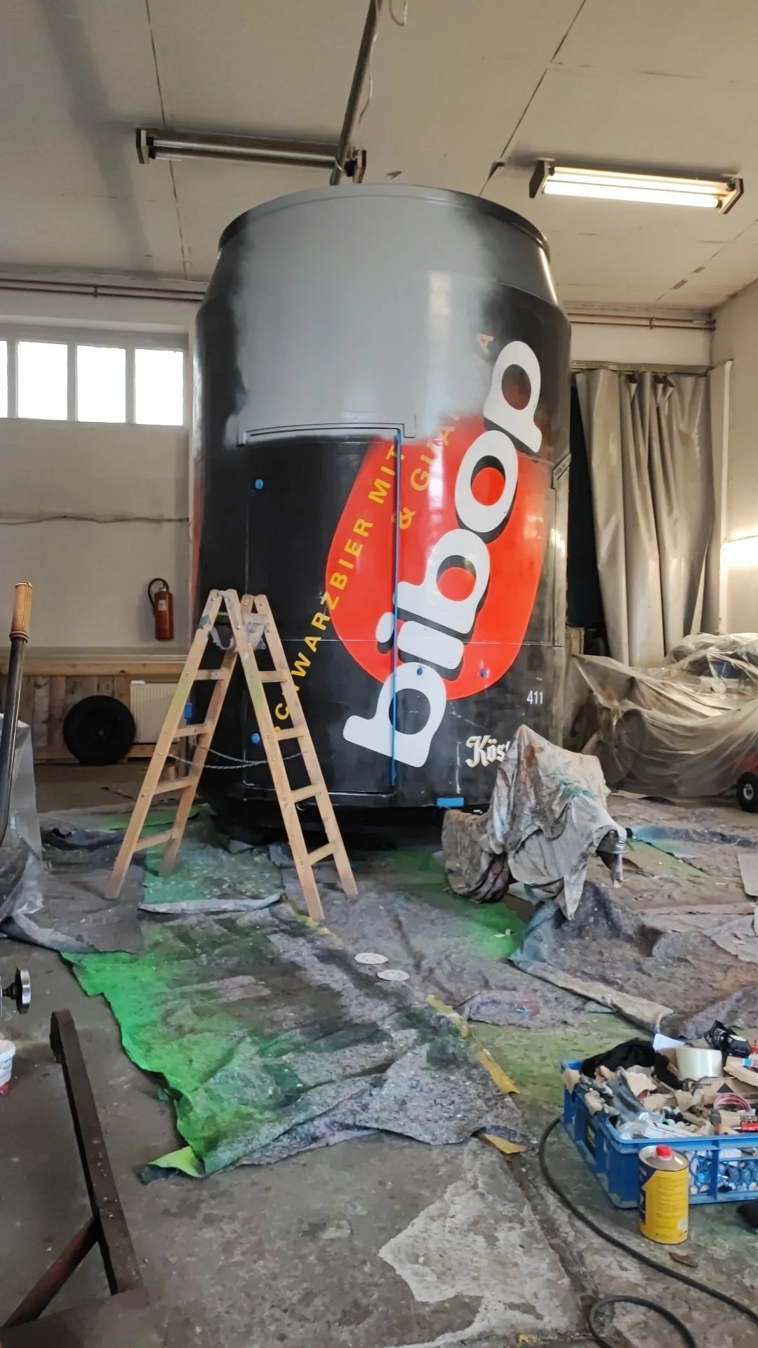 Ein großer umgestalteter Getränkecontainer mit einem Coca-Cola-Logo, in einem Werkstatt- oder Lagerraum, umgeben von Werkzeugen, Leiter und Abdeckmaterialien.