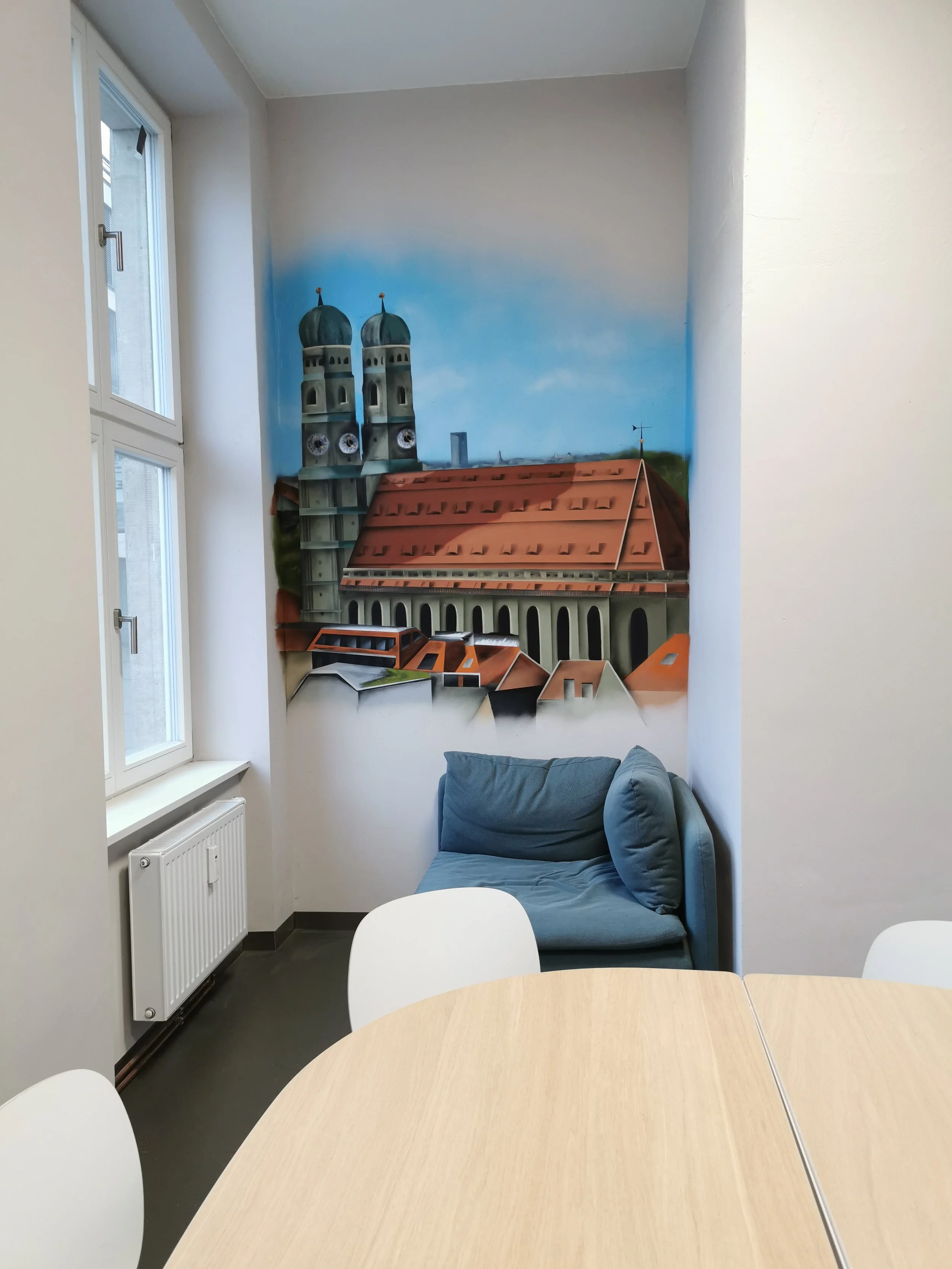 Zimmer mit einem Fenster, einer blauen Couch in einer Ecke, einem runden Holztisch und einer Wandmalerei eines Gebäudes mit Türmen, blauen Kuppeln und roten Dächern.