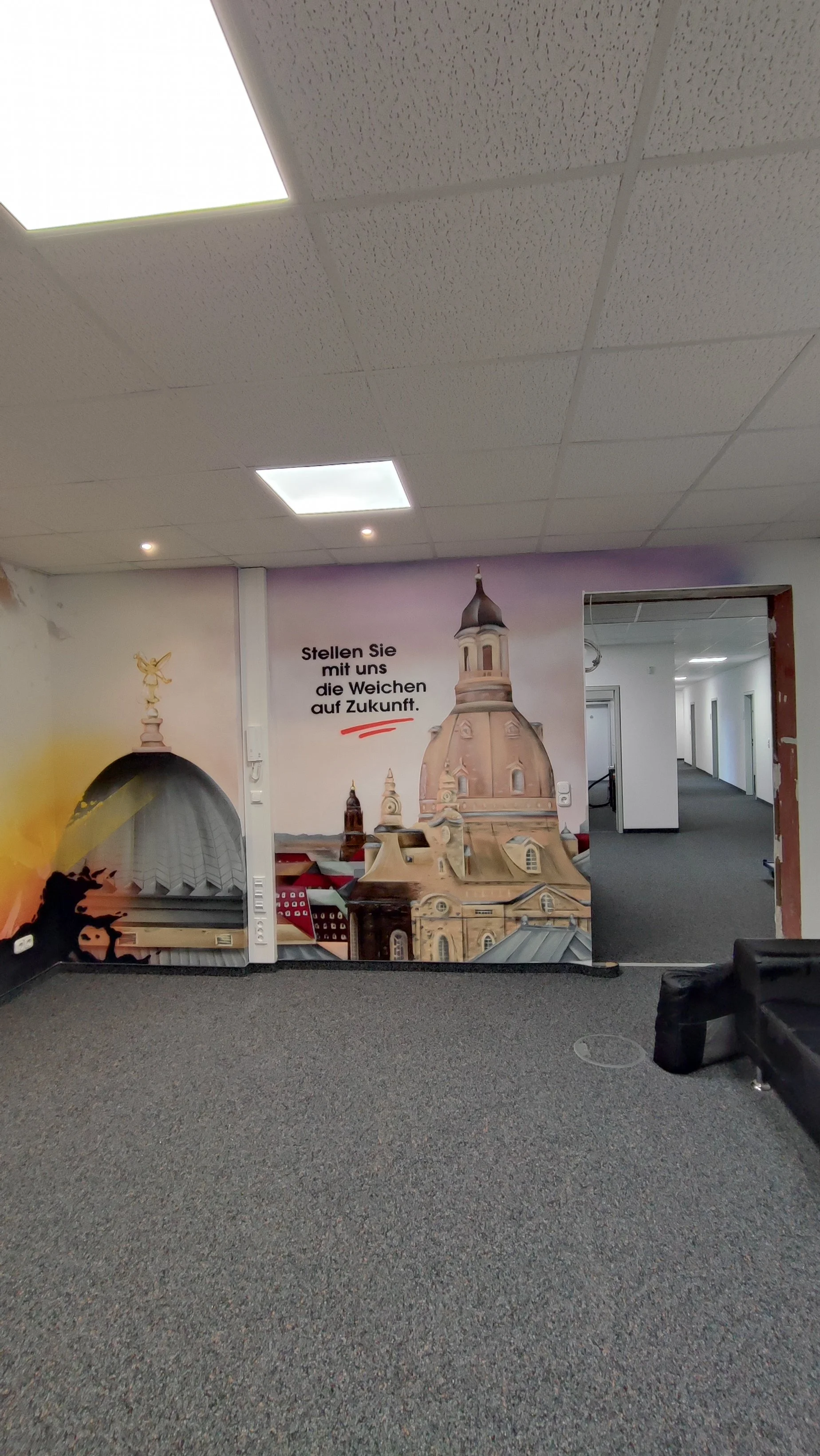 Wand mit Wandbild, das eine Stadtsilhouette mit Kirchen und Türmen zeigt, sowie den deutschen Text 'Stellen Sie mit uns die Weichen auf Zukunft'.