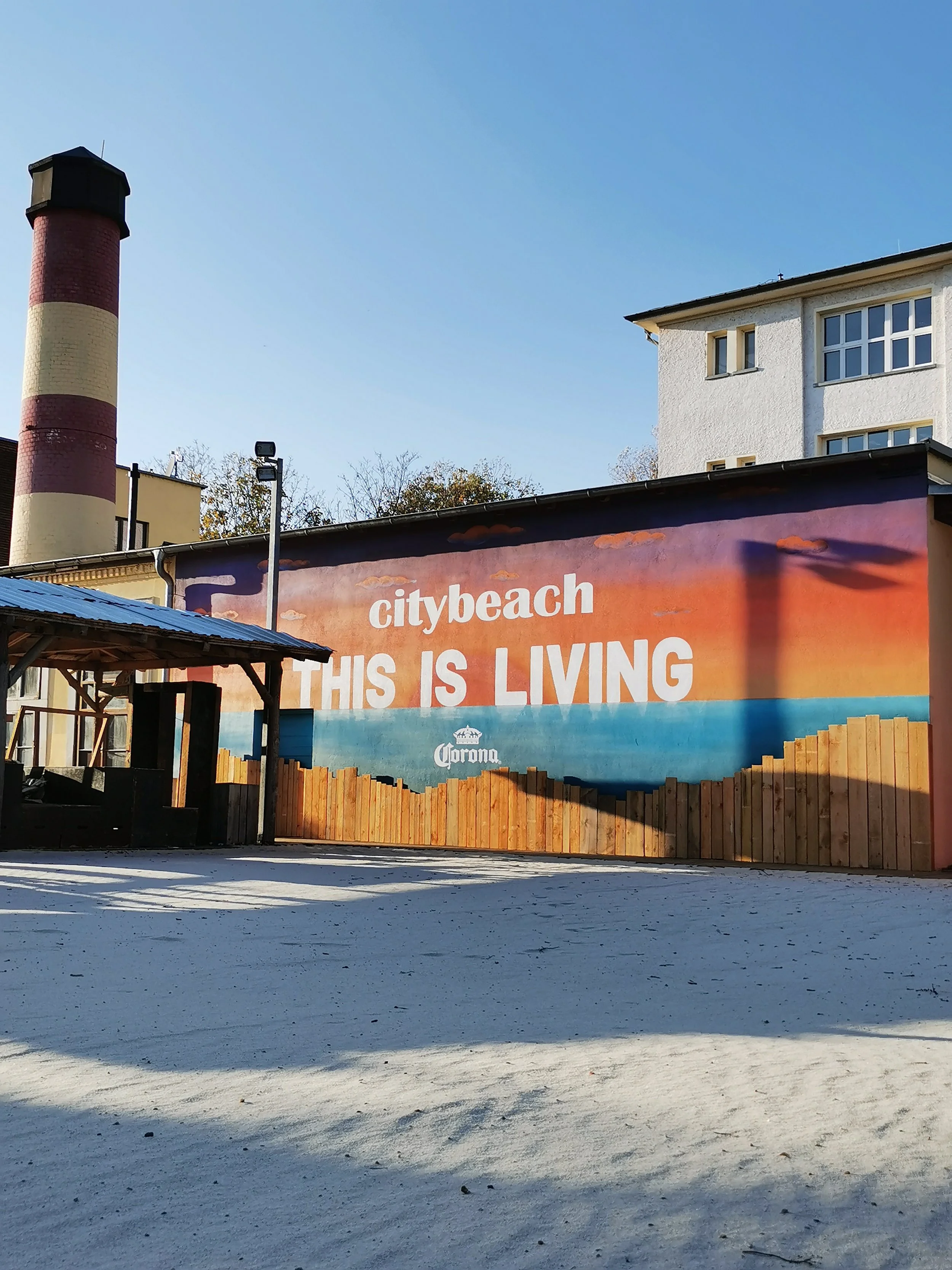 Wand mit Slogan 'citybeach THIS IS LIVING' und Logo von Corona, im Hintergrund Gebäude und ein Schornstein, außenbereich bei Sonnenuntergang.