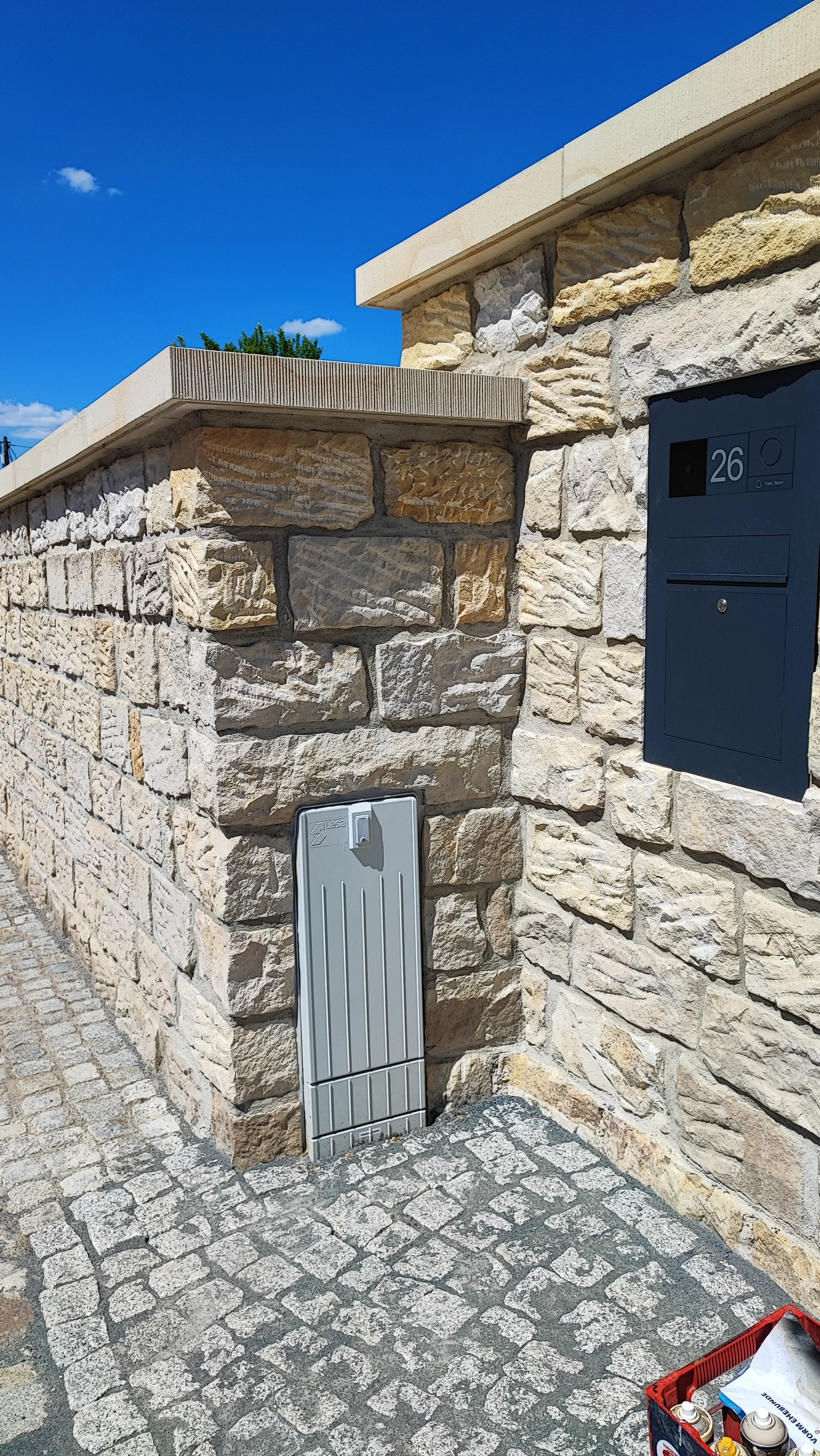 Straße mit Steinmauer, Briefkasten, Zählerkasten, kleine Kiste mit Farben und Werkzeugen, blauer Himmel mit wenigen Wolken.