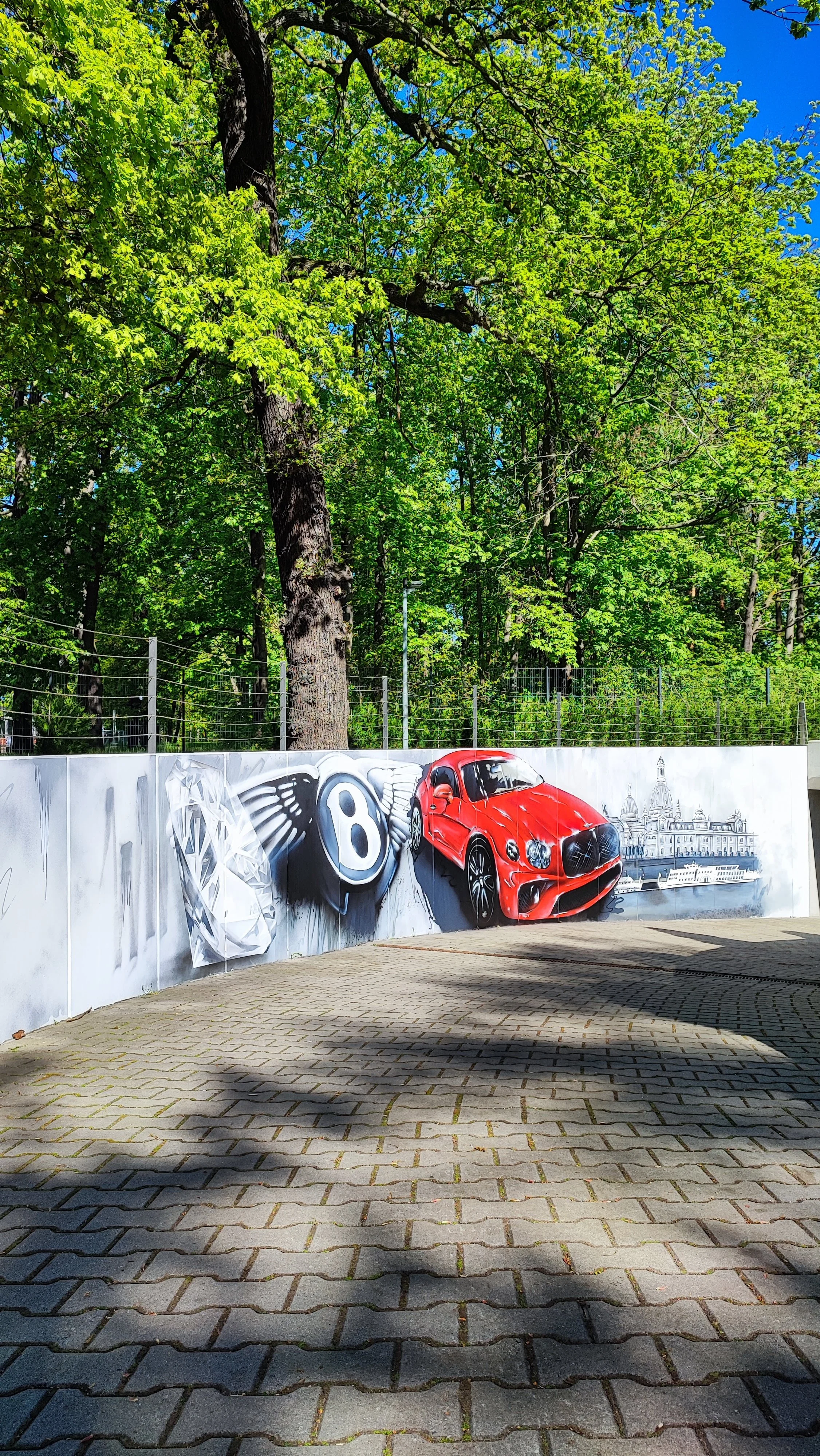 Straßenmalerei an einer Wand mit einem Diamanten, einem Bentley-Logo, einem roten Sportwagen und einer Stadtansicht mit einem Fluss und einer Kirche.