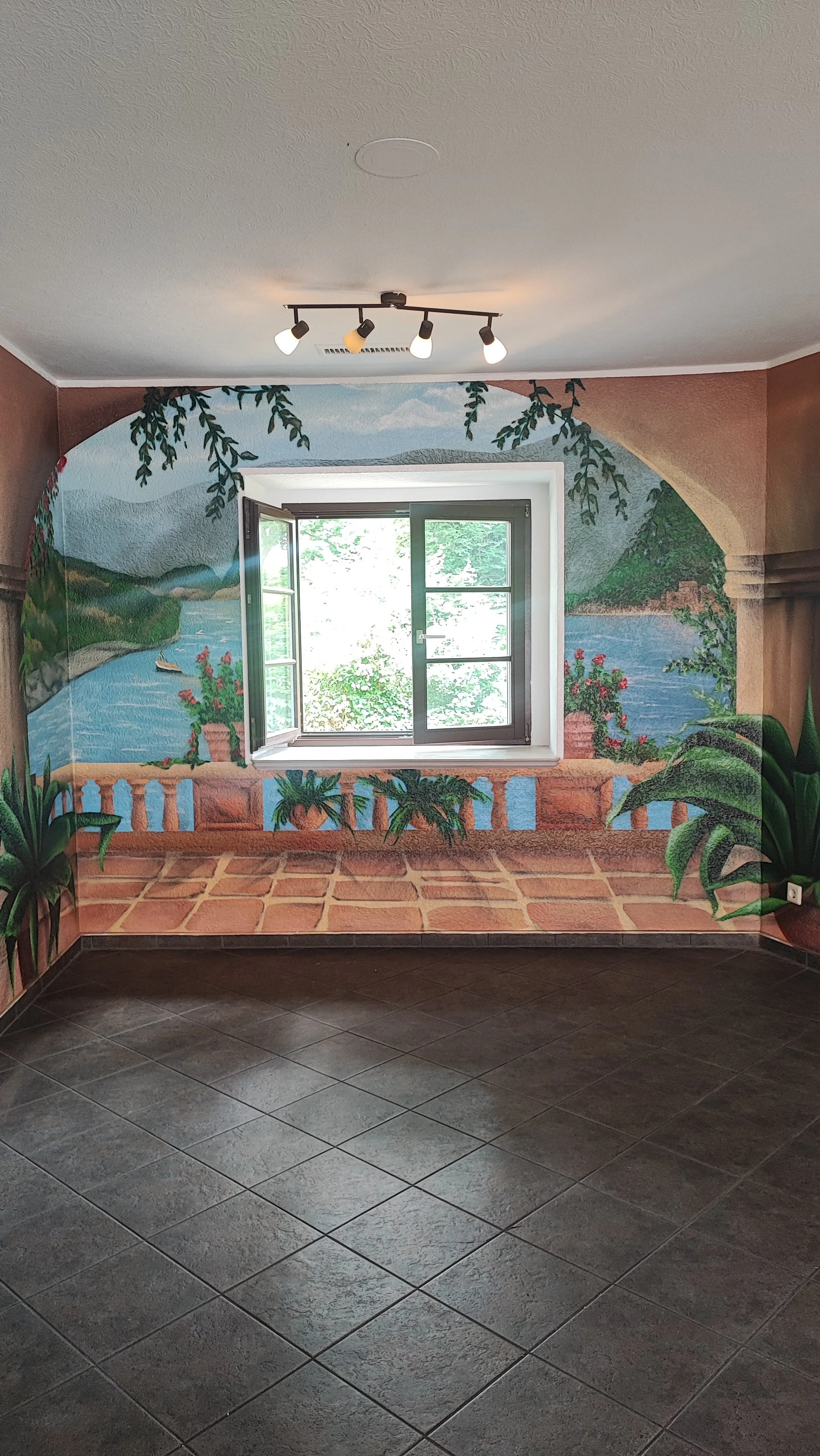 Zimmer mit Wandmalerei einer Balkonansicht mit Fluss, Landschaft und Pflanzen, eine offene Fensteröffnung, Fliesenboden, Deckenleuchte.