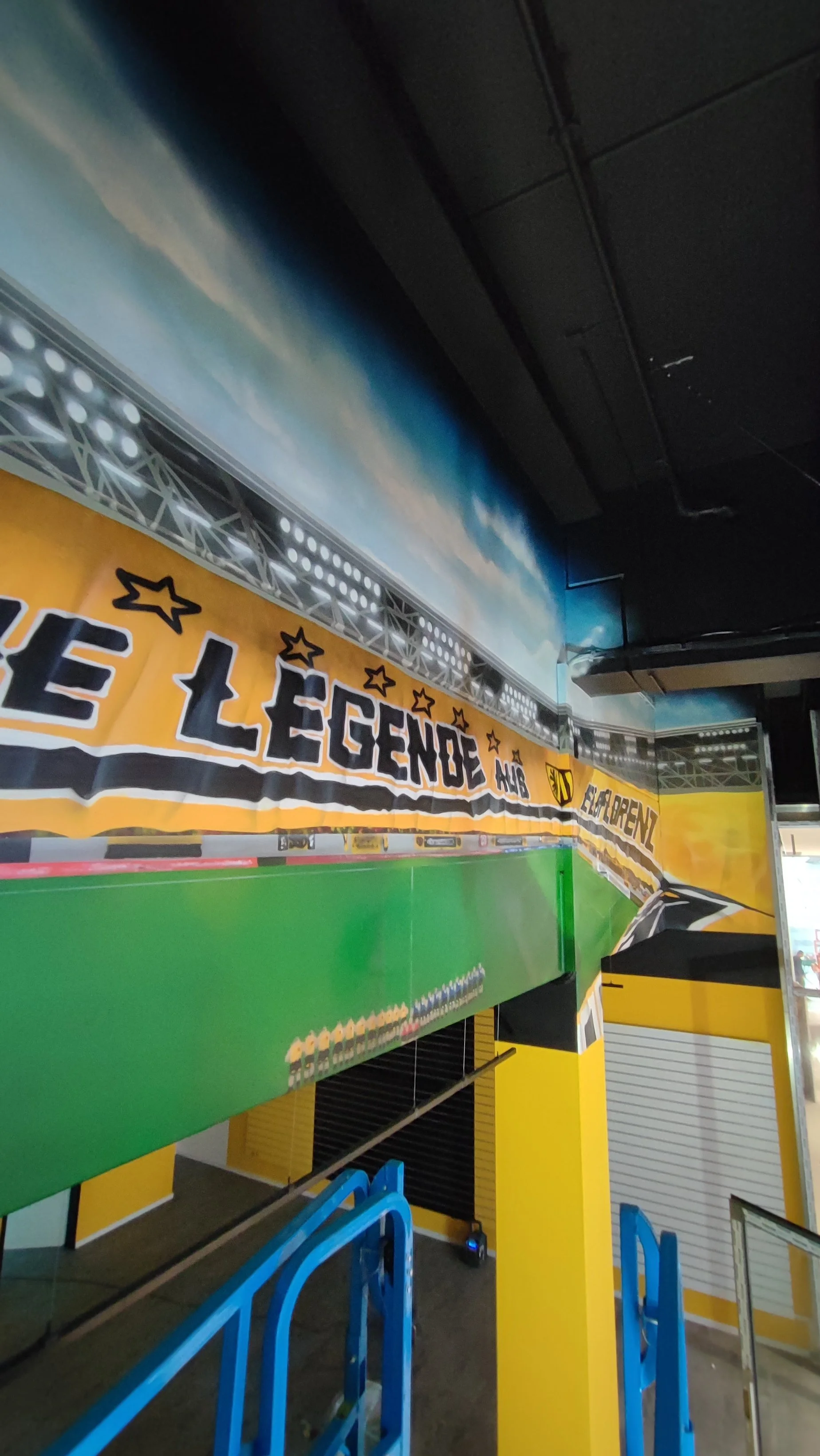 Blick auf eine Indoor-Fußballbahn mit gelbem Banner, das den Text "THE LEGENDS AL4" und einen Aachener StadtWappen zeigt.