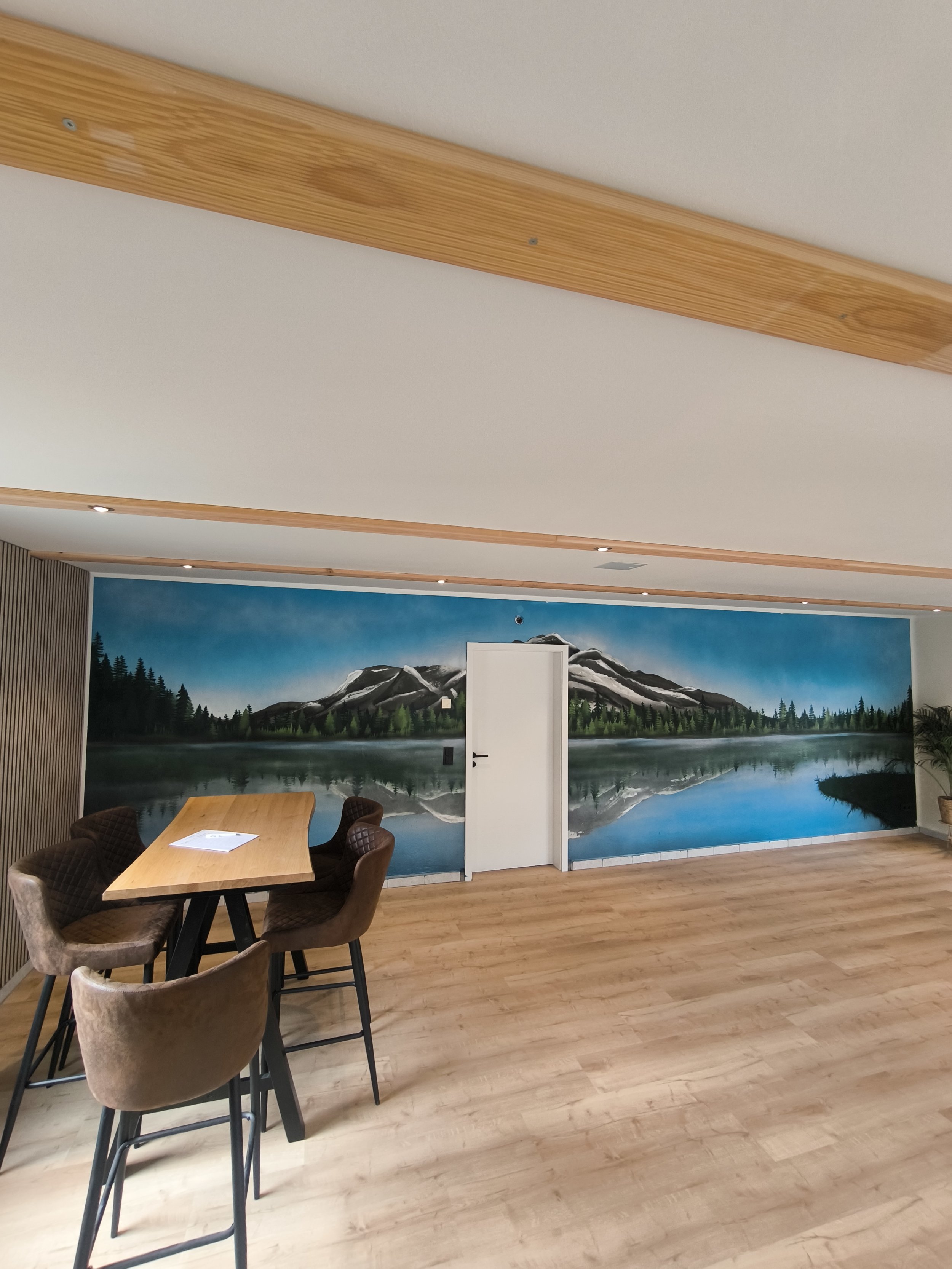 Ein Raum mit einem Tisch und vier Stühlen vor einer Wandmalerei eines Sees mit Bergen und Wald im Hintergrund.