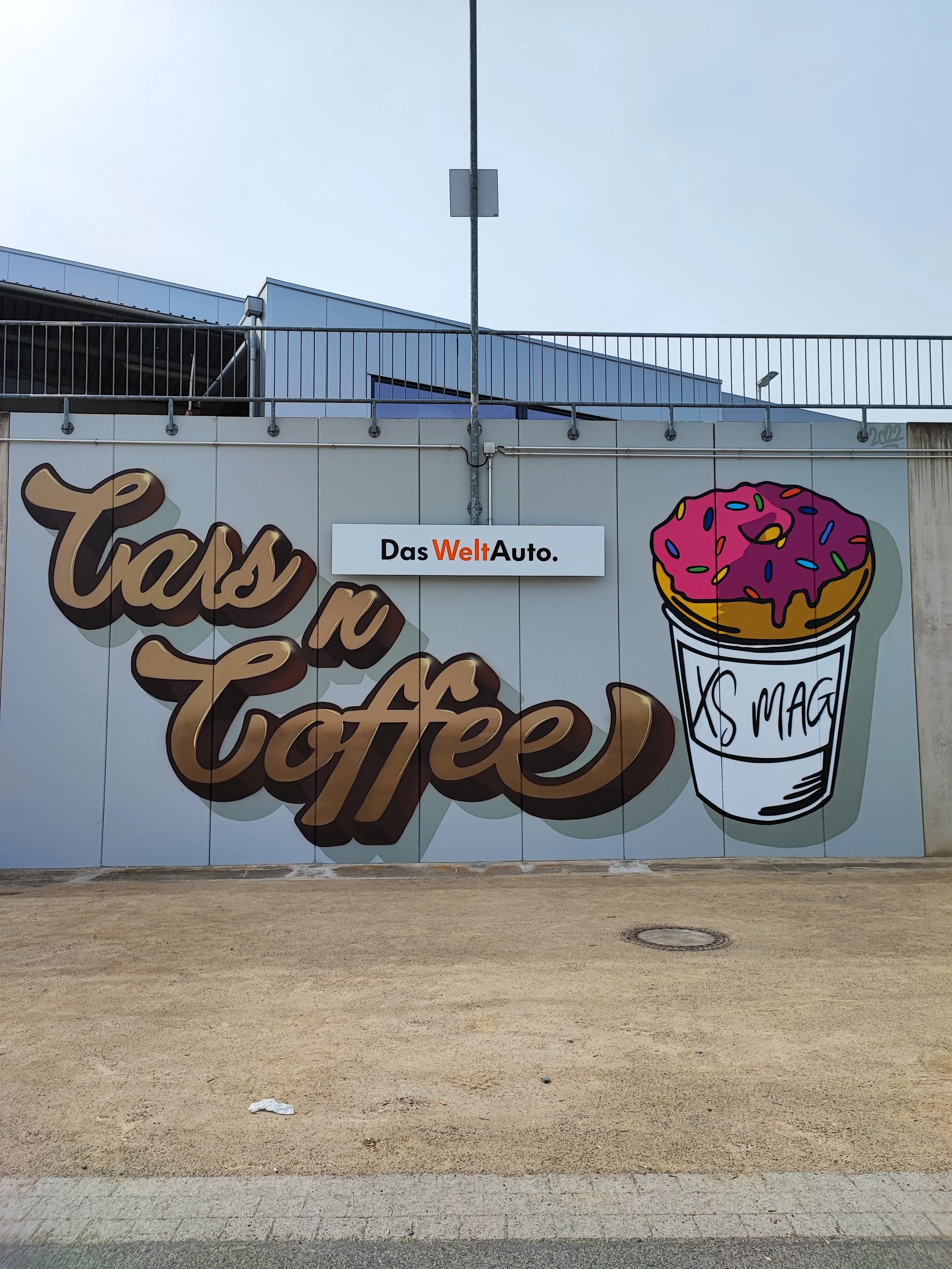 Werbetafel mit der Aufschrift "Tass n Coffee" und einem großen Bild eines Donuts mit pinkem Zuckerguss und bunten Streuseln sowie einem Kaffee-Becher mit der Aufschrift "XS MAG". Ein weißes Schild mit dem Text "Das WeltAuto" ist darüber angebracht.