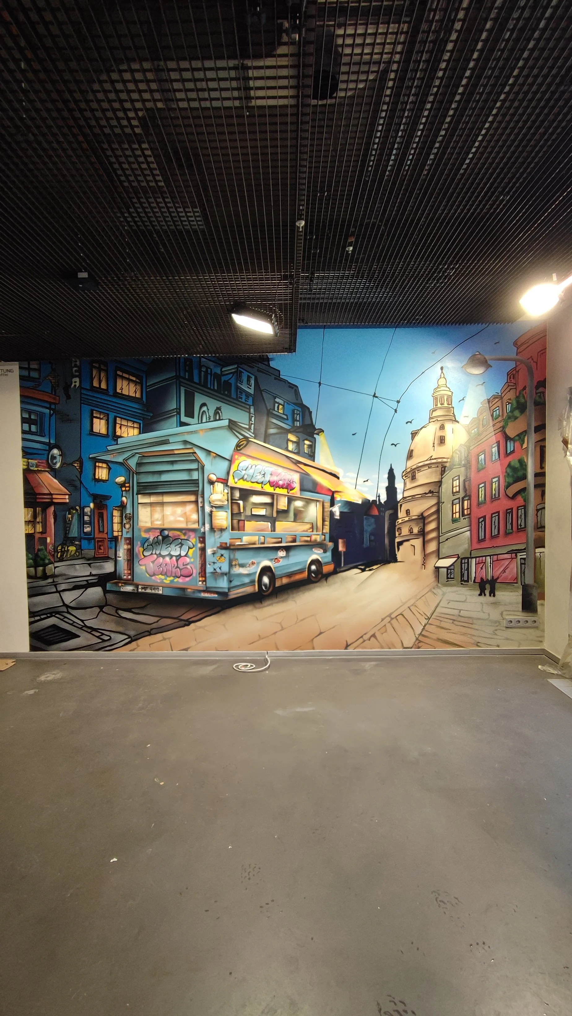 Street-Art Wand Gemälde eines Straßenkiosk auf einer Stadtstraße, umgeben von bunten Gebäuden und einer Kuppel, in lebendigen Farben, in einem Innenraum mit dunklem Gitterdecke