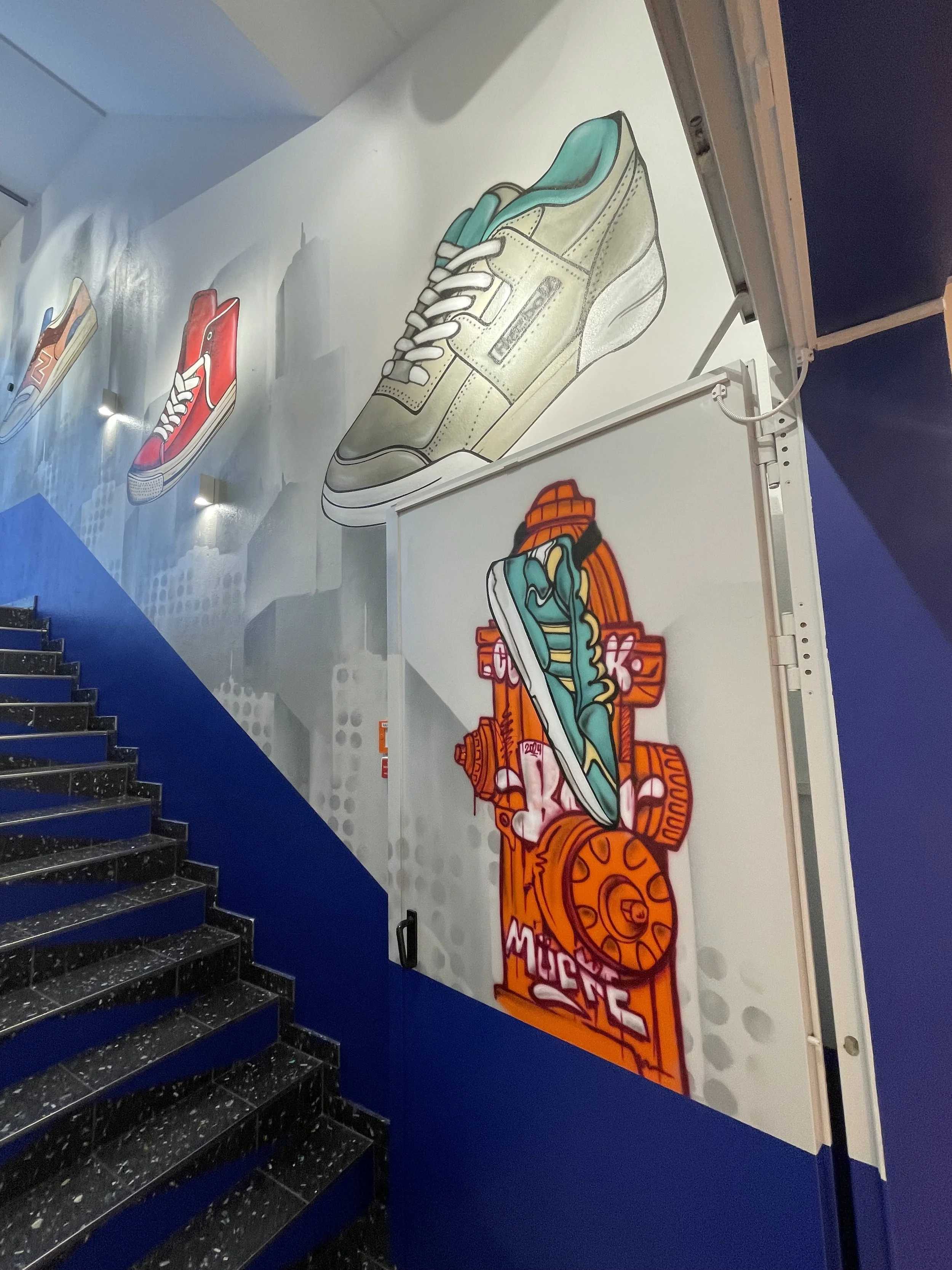 Graffiti an der Wand mit großen Bildern verschiedener Turnschuhe und einem rot-orangefarbenen Graffiti-Tags, das Schuhe und eine Uhr zeigt.