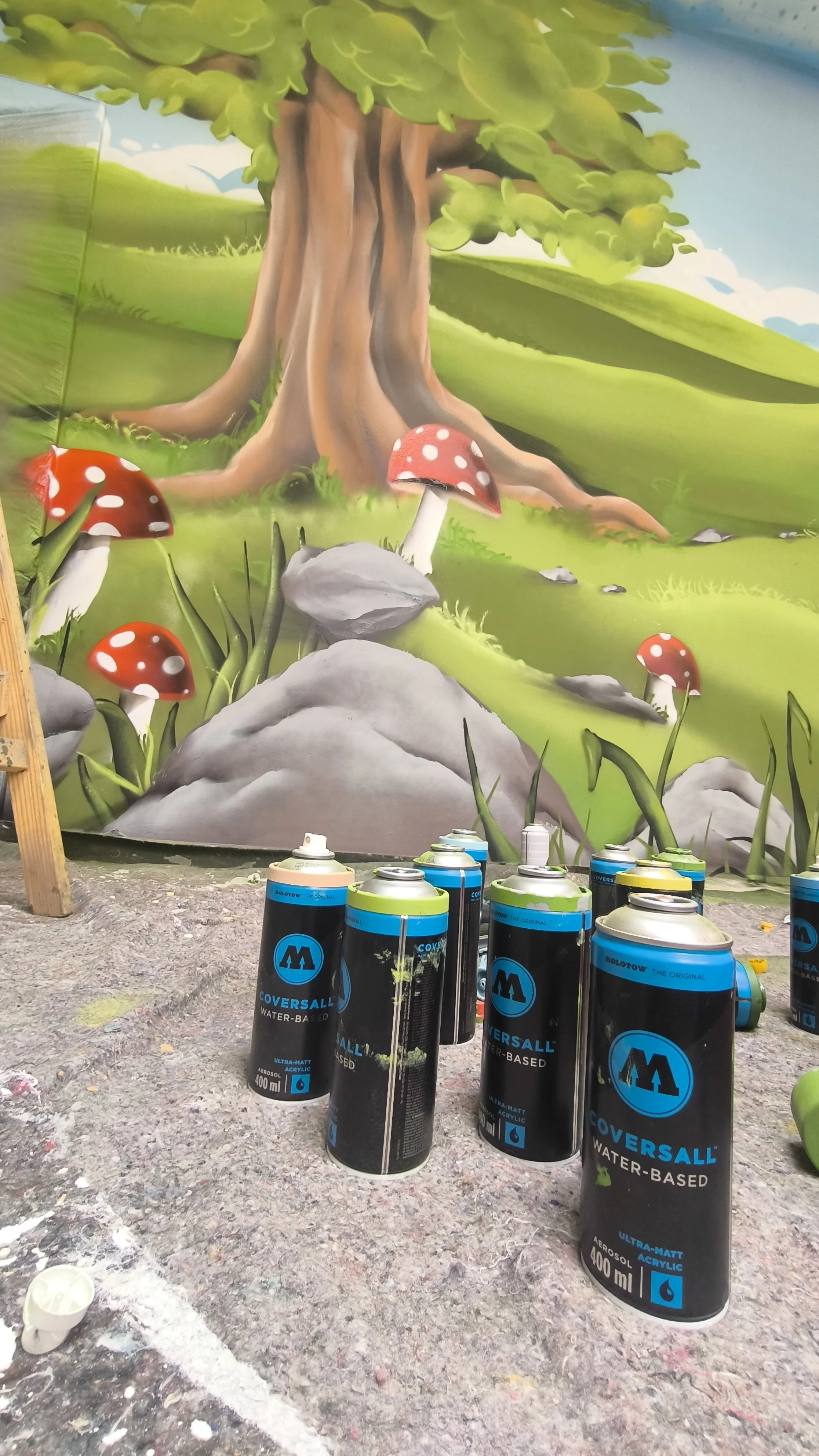 Mural mit einem großen Baum, roten Pilzen mit weißen Punkten und bunten Gras. Mehrere Spraydosen vor der Wand.