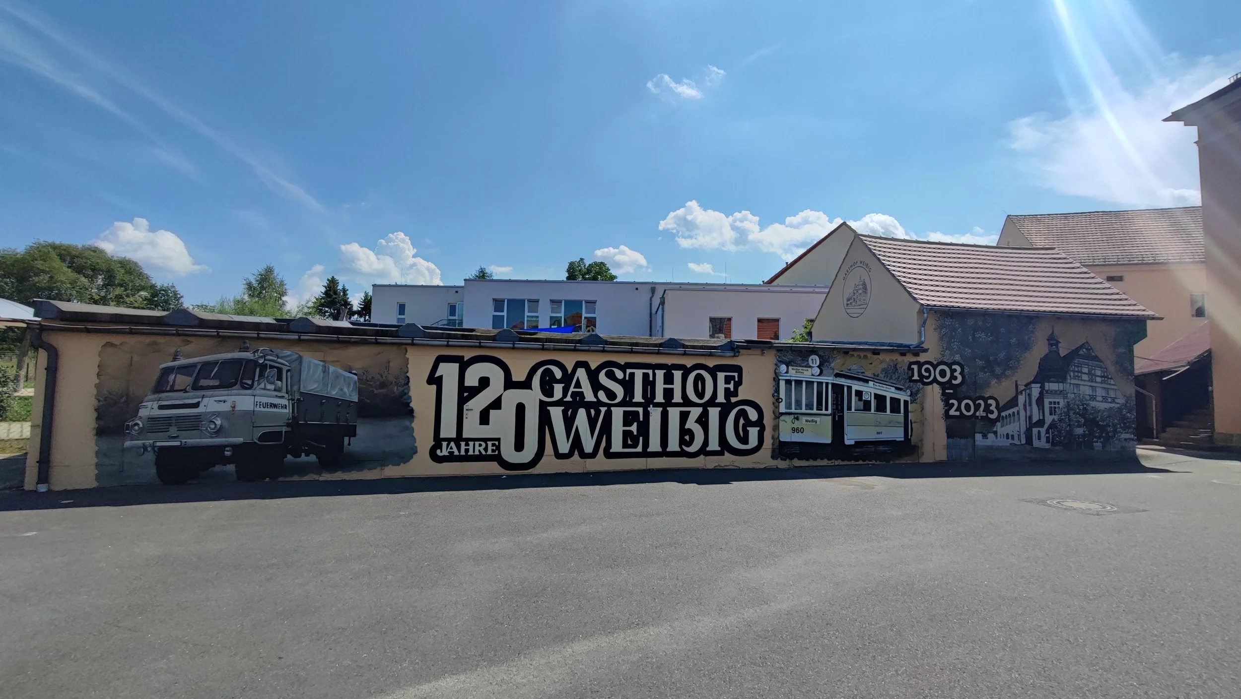 Wand bemalt mit historischem Gasthof und Straßenbahn, die 120 Jahre feiert, mit Texten und Bildern, darunter ein Feuerwehrauto, der Name 'Gasthof Weißig', das Gründungsjahr 1903 und das Jubiläumsjahr 2023 in Deutschland.