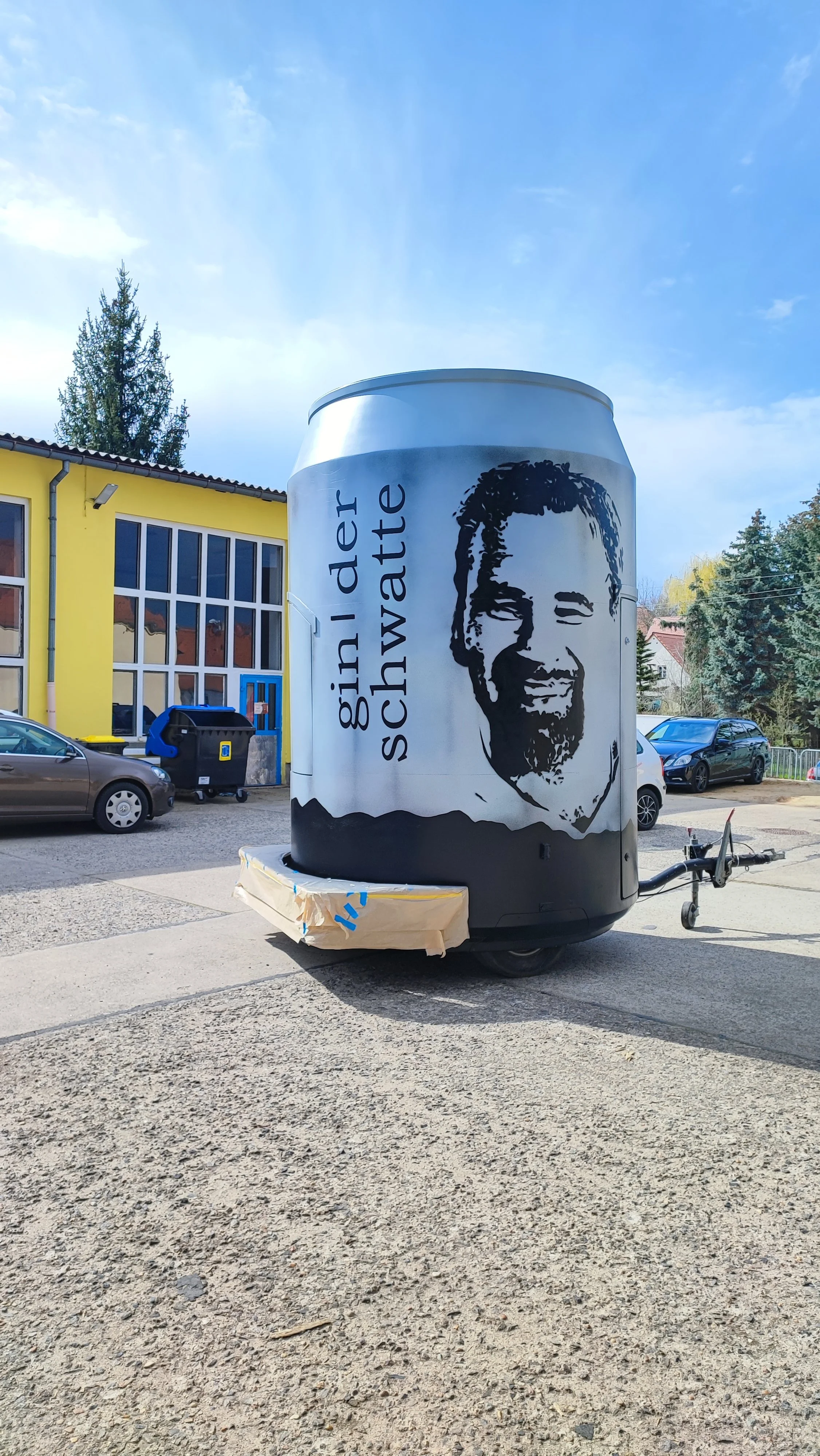 Ein Rollcontainer, gestaltet wie eine Getränkedose, mit einem Porträt und dem Text »GIRL ADER Schwarzwa« auf dem Gelände eines Parkplatzes. Im Hintergrund sind Autos und ein gelbes Gebäude zu sehen.