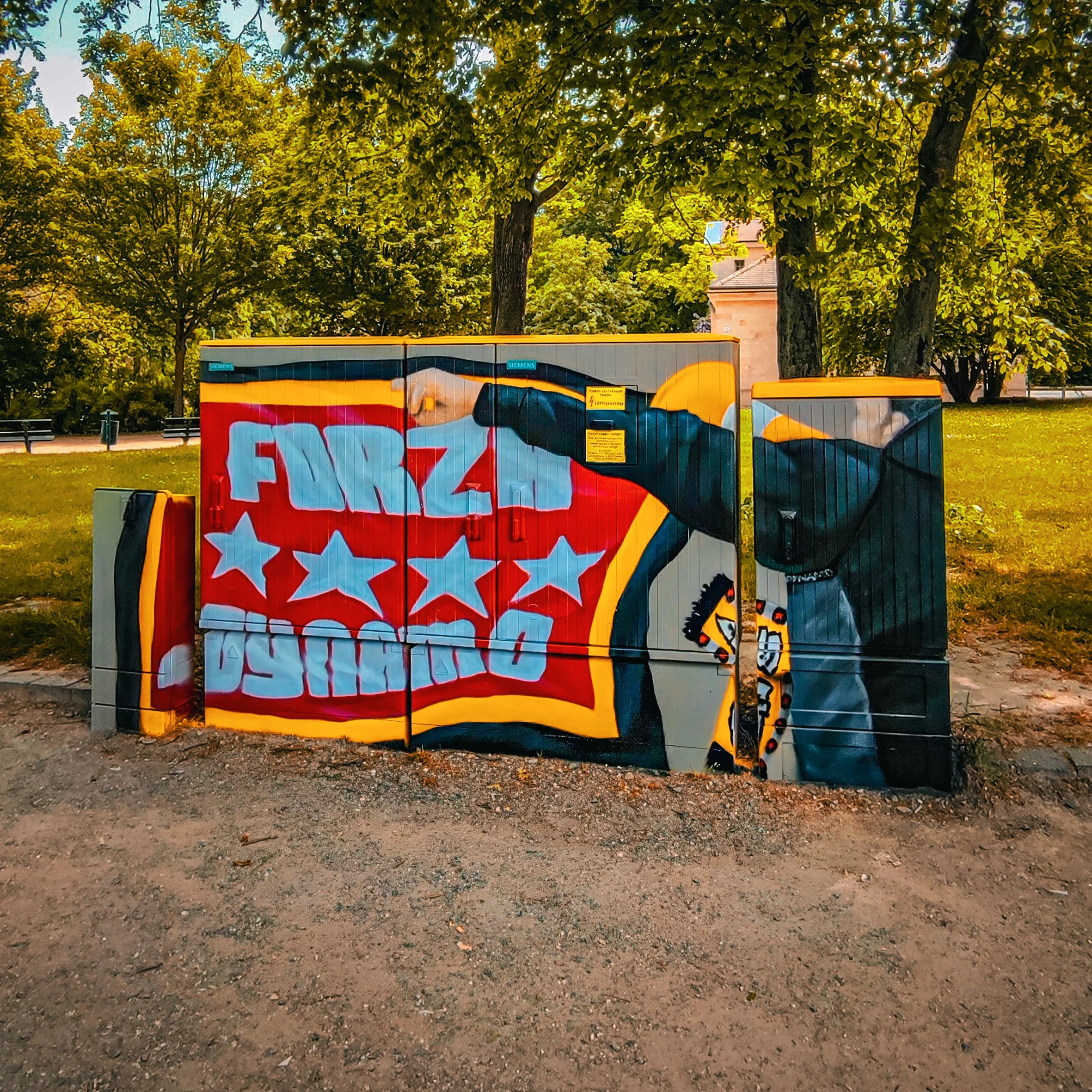 Graffiti-Kunstwerk auf einer Stromkasten in einem Park mit grünen Bäumen im Hintergrund. Das Bild zeigt eine Figur, die einen Baseballschläger hält, sowie Text und Sterne im Graffiti-Stil.