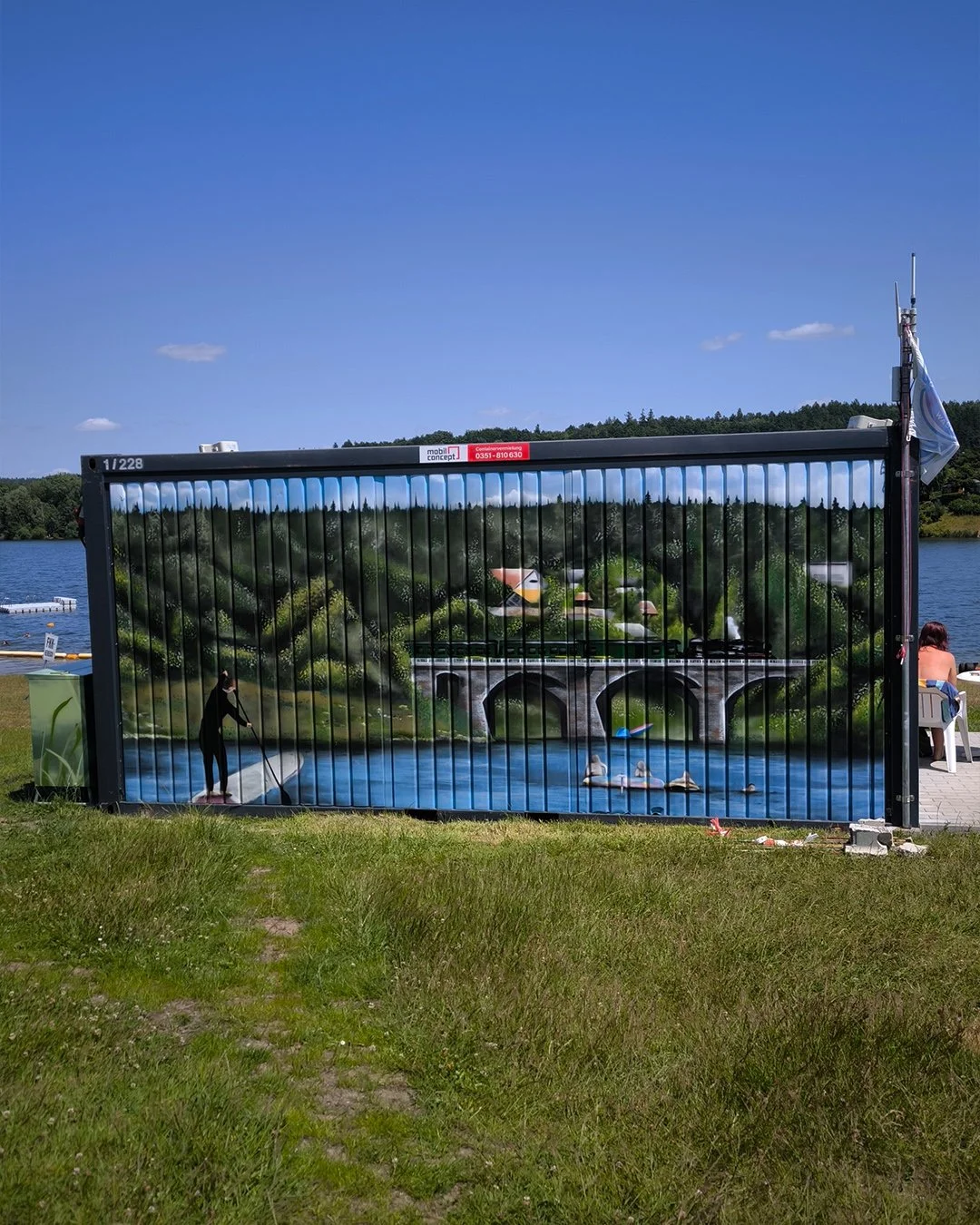 Ein aufgeblasenes Werbeplakat mit einer malerischen Landschaft, das einen Fluss, eine Brücke, Bäume und Häuser zeigt. Das Plakat befindet sich in einem Zaun am Wasser, im Hintergrund sind Wasser, Bäume und Himmel sichtbar.