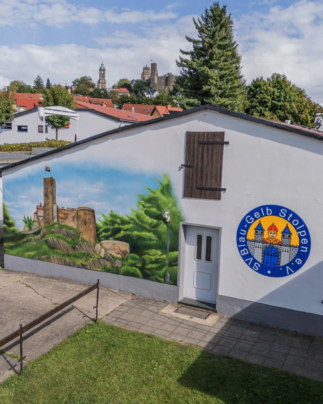 Wandbild an einer weißen Hauswand mit Gemälde einer Burg auf einem Hügel, umgeben von Bäumen, und einem blauen Himmel im Hintergrund.