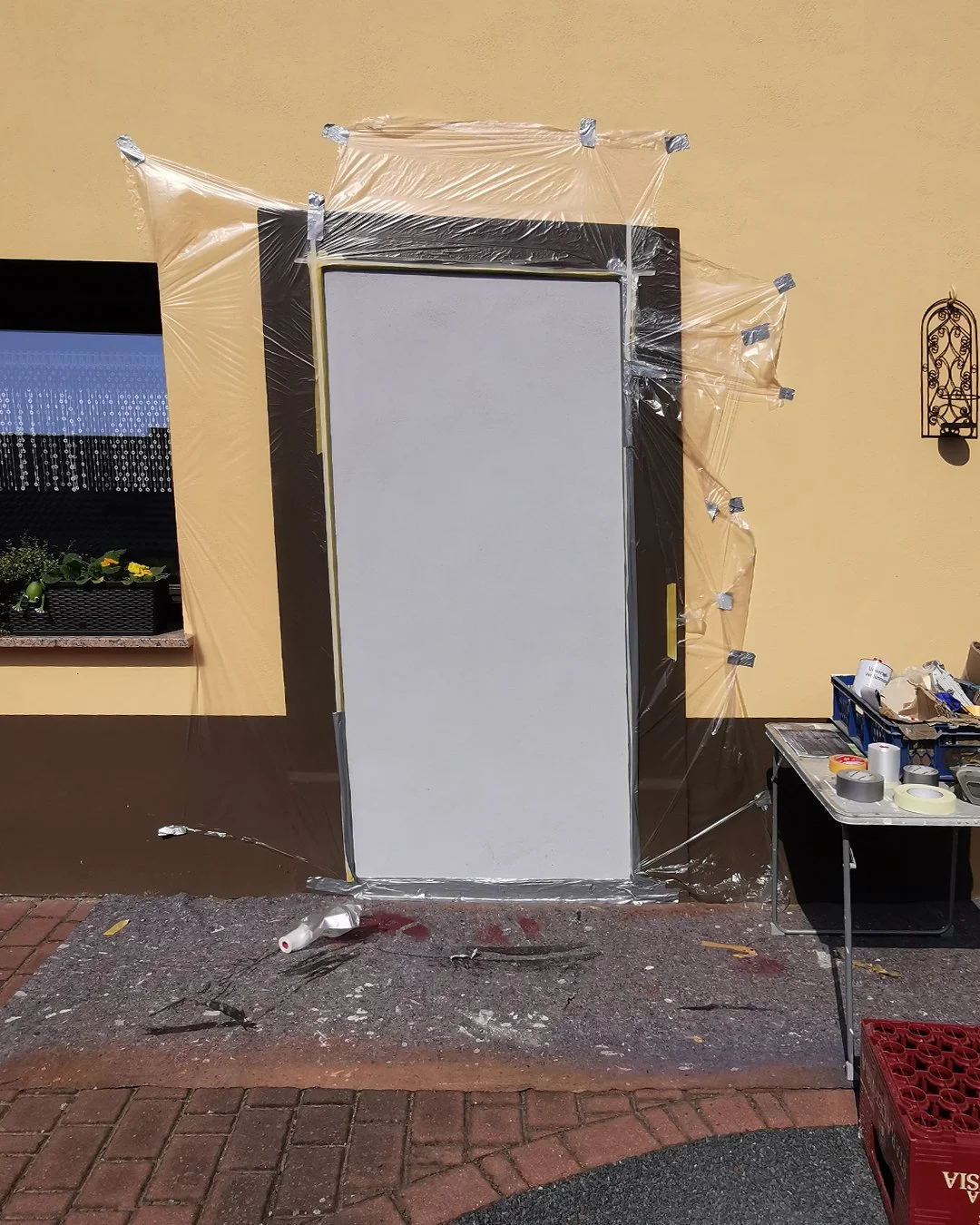 Festlich dekorierte weiße Tür vor gelbem Haus mit Fenster und Pflanzkasten, umrahmt von transparentem Plastik und Klebeband, mit Werkbank und Müllcontainer auf Gehweg.