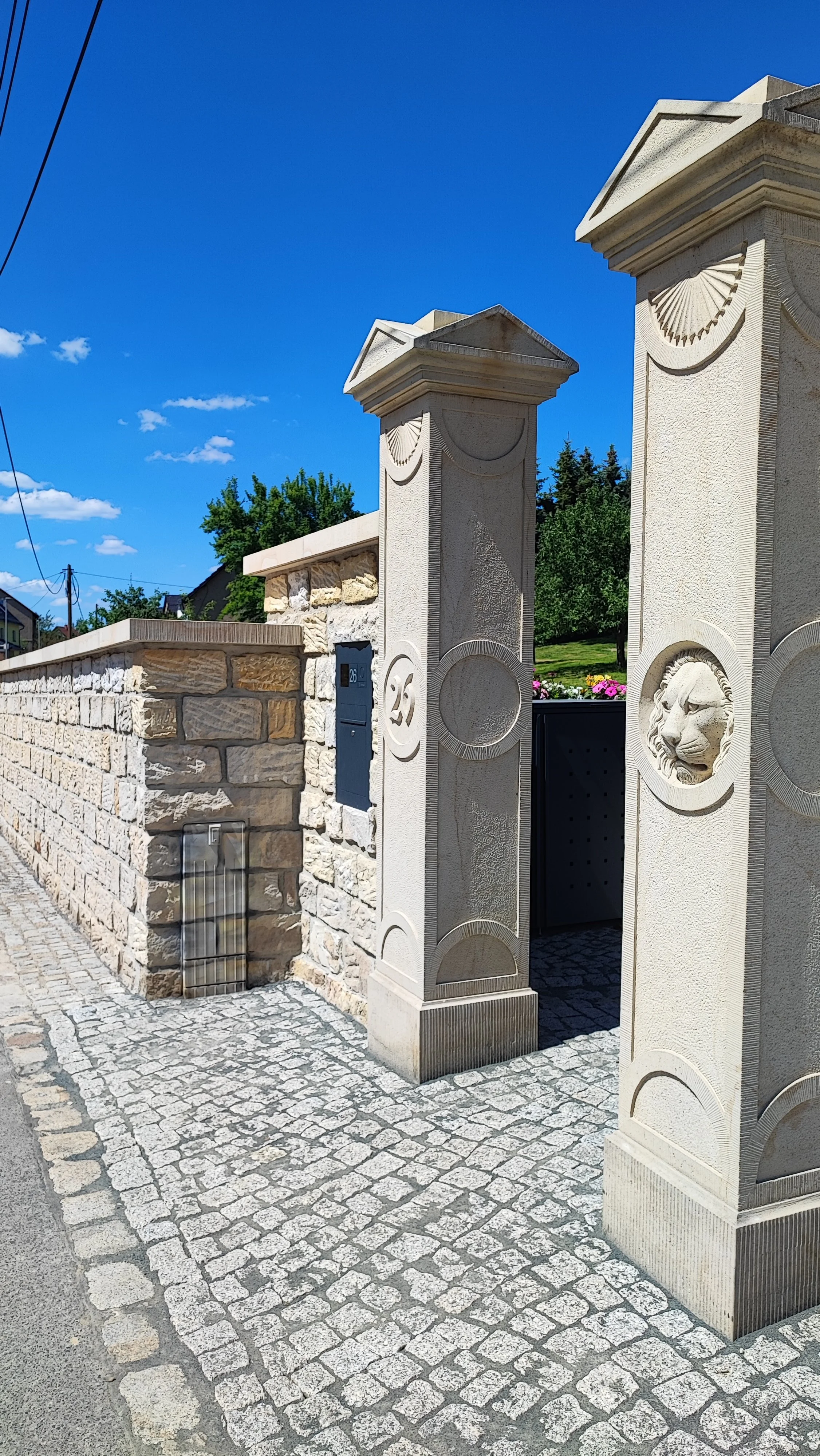 Ein Holzzaun mit Steinmauer, zwei Säulen mit Löwenkopfdekoration, blauer Himmel und grüne Bäume im Hintergrund.