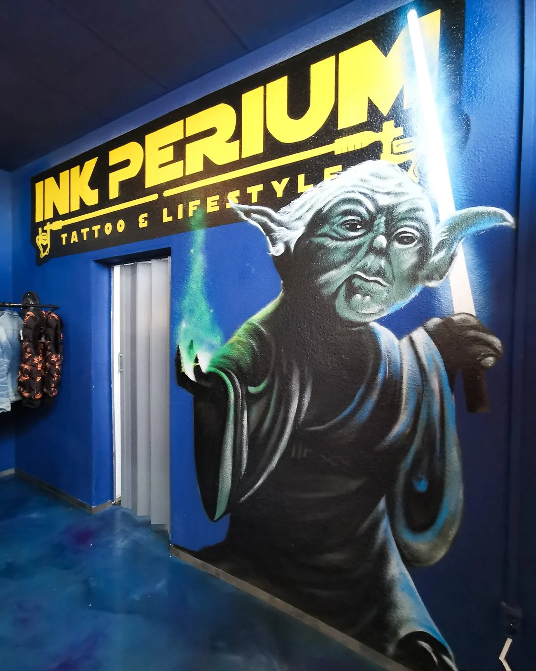 Graffiti an einer blauen Wand mit der Aufschrift 'INK PERIUM TATTOO & LIFESTYLE' und einer Darstellung von Yoda aus Star Wars, der auf der Wand erscheint.