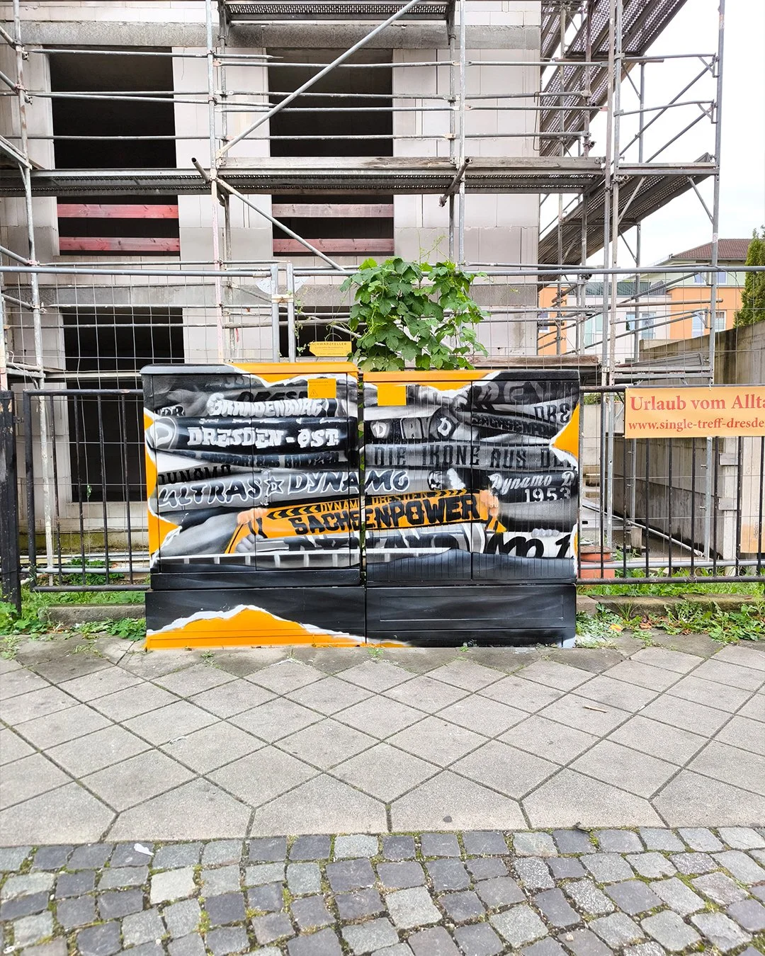 Ein Bauzaun mit Graffiti in einer städtischen Umgebung, im Hintergrund ein Gebäude mit Gerüsten und grüner Pflanzen, rechts ein gelber Kasten sowie ein orangefarbenes Schild mit Website-URL.
