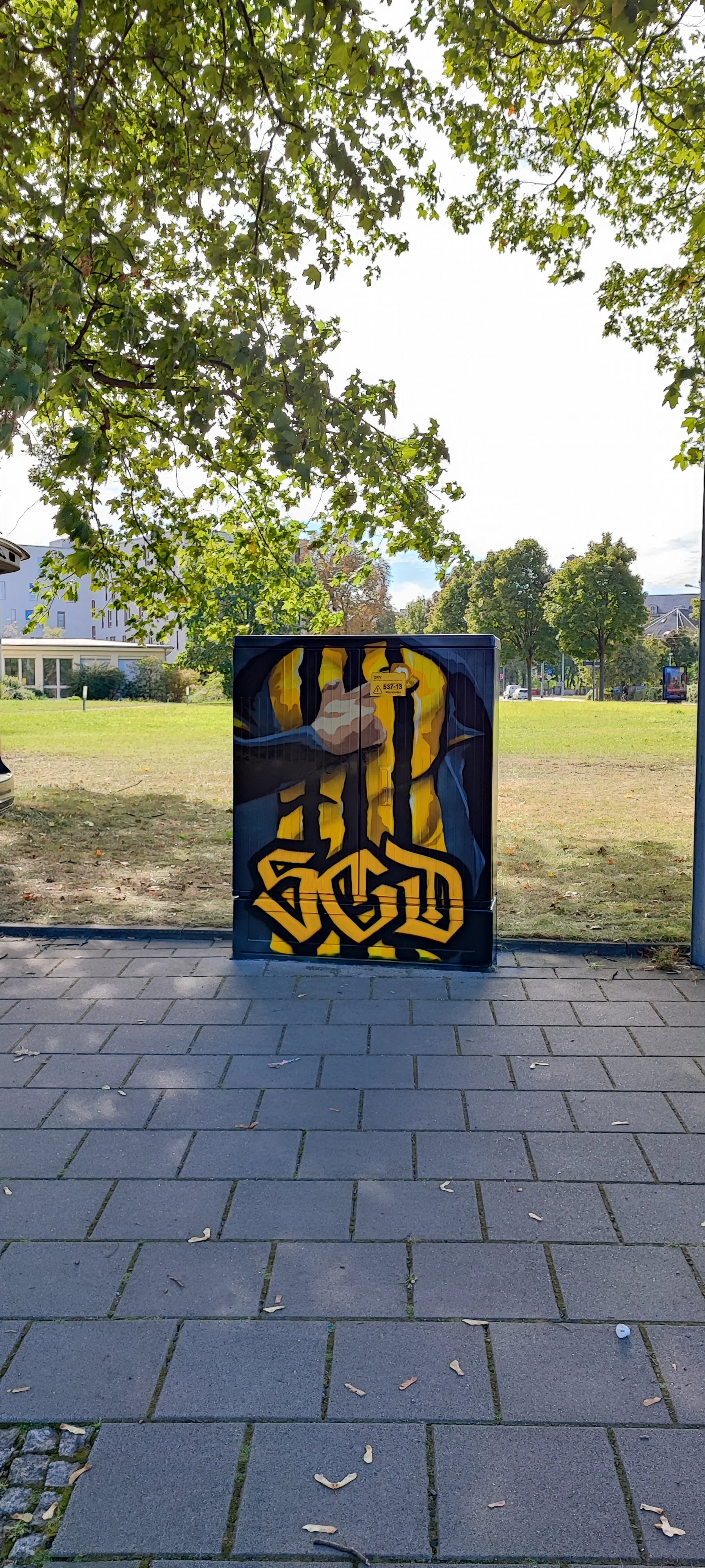 Graffiti-Kunst auf einer Stromkasten, das eine muskulöse Figur zeigt, die einen Kraftakt zwischen zwei Kabeln ausführt, mit grünen Bäumen im Hintergrund.