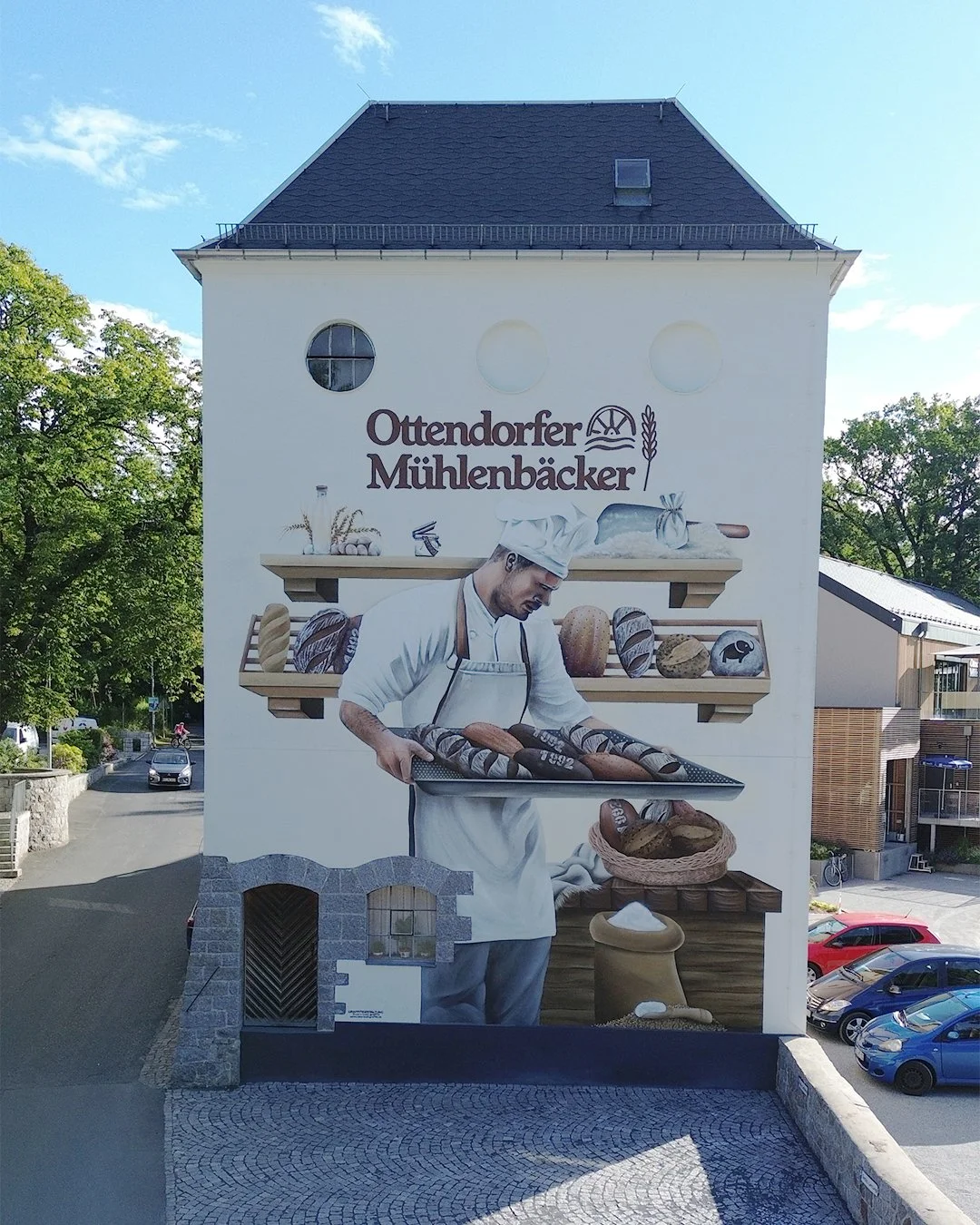 Wandbild eines Bäckers, der auf einer Tafel frisches Brot präsentiert. Der Schriftzug lautet "Ottendorfer Mühlenbäcker". Professionelle Graffitigestaltung in Dresden: individuelle Wandgestaltung, Auftragsgraffiti sowie kreative Graffiti-Workshops und
