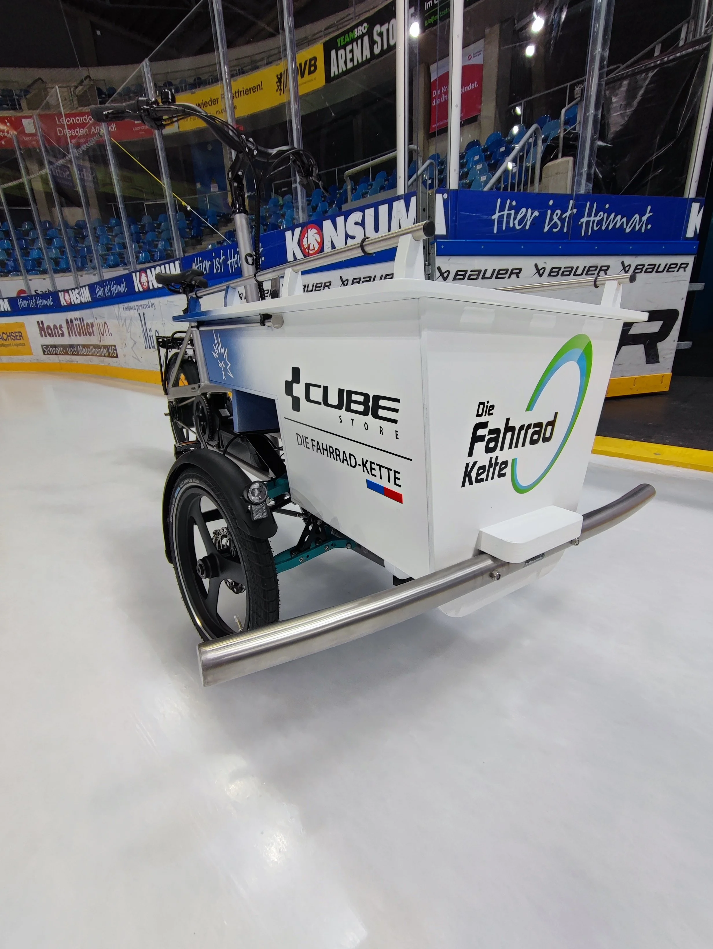 Ein innovatives elektrisches Fahrrad-Rikschafahrzeug auf einer Eisbahn, ausgestattet mit einem Anhänger für den Transport, mit sichtbaren Logos und Text in Deutsch auf den Seiten. Professionelle Graffitigestaltung in Dresden: individuelle Wandgestalt