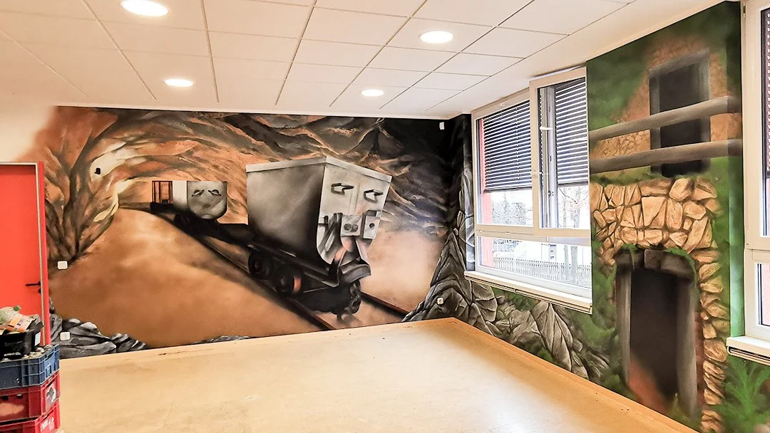 Wandgemälde zeigt einen Bergwerkszug in einer Galerie, mit Felsen und grünen Pflanzen.