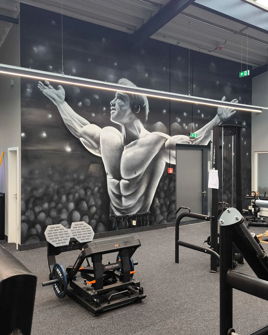 Schwarz-weiß Wandbemalung in Fitnessstudio, zeigt muskulösen Mann mit ausgebreiteten Armen vor einer Bühne, mit Fitnessgeräten im Vordergrund.