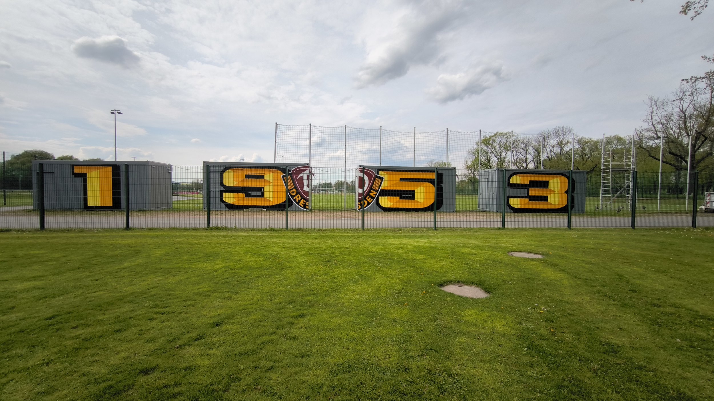 Schild mit großen gelben Nummern 1, 3, 5, und 3 auf einem Sportplatz, umgeben von einem Zaun, mit Bäumen und Wolken am Himmel im Hintergrund.