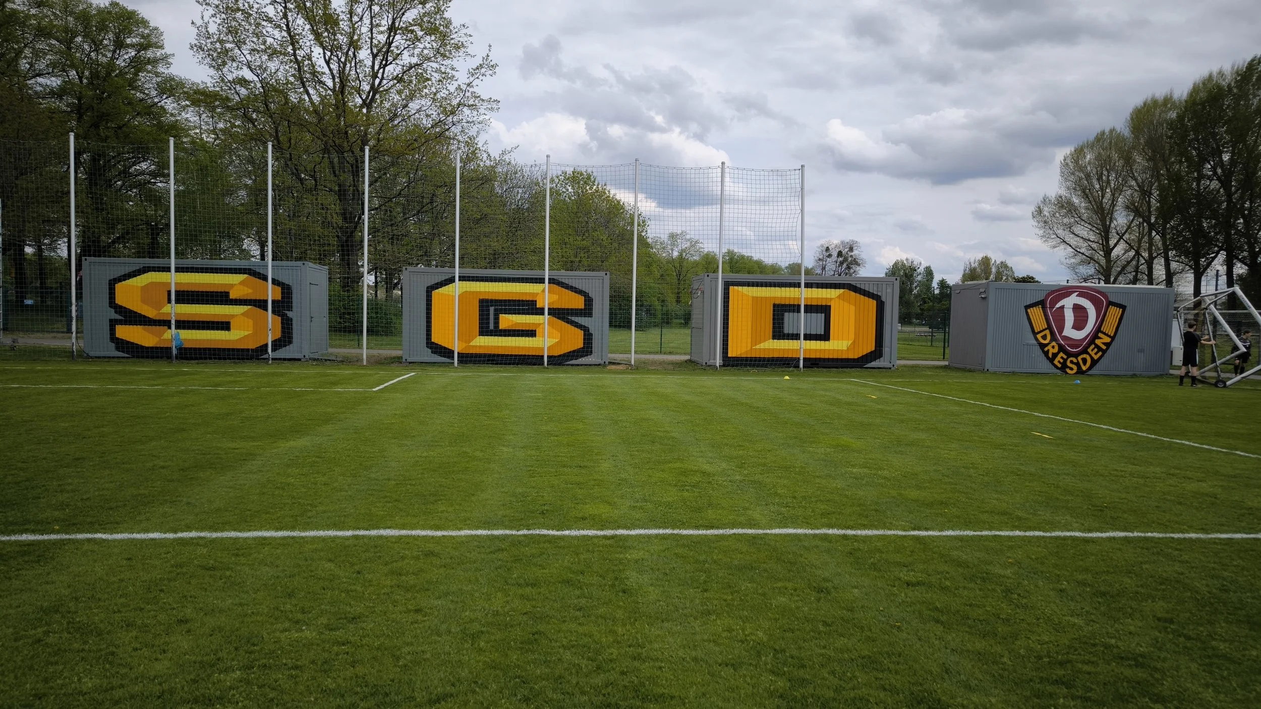 Auf dem Fußballfeld sind Container mit den Farben und dem Logo des FC Dynamo Dresden bemalt, die die Buchstaben S, G, D bilden. Der Himmel ist bewölkt, und im Hintergrund stehen Bäume.