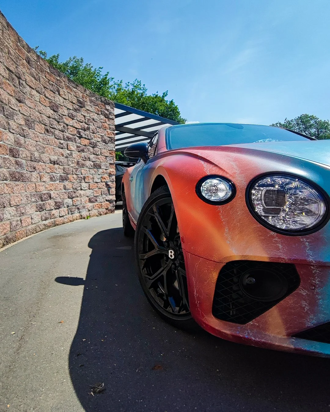Sportwagen mit buntem Lack, schwarze Felgen, nahe an einer Steinmauer im Freien unter blauem Himmel.