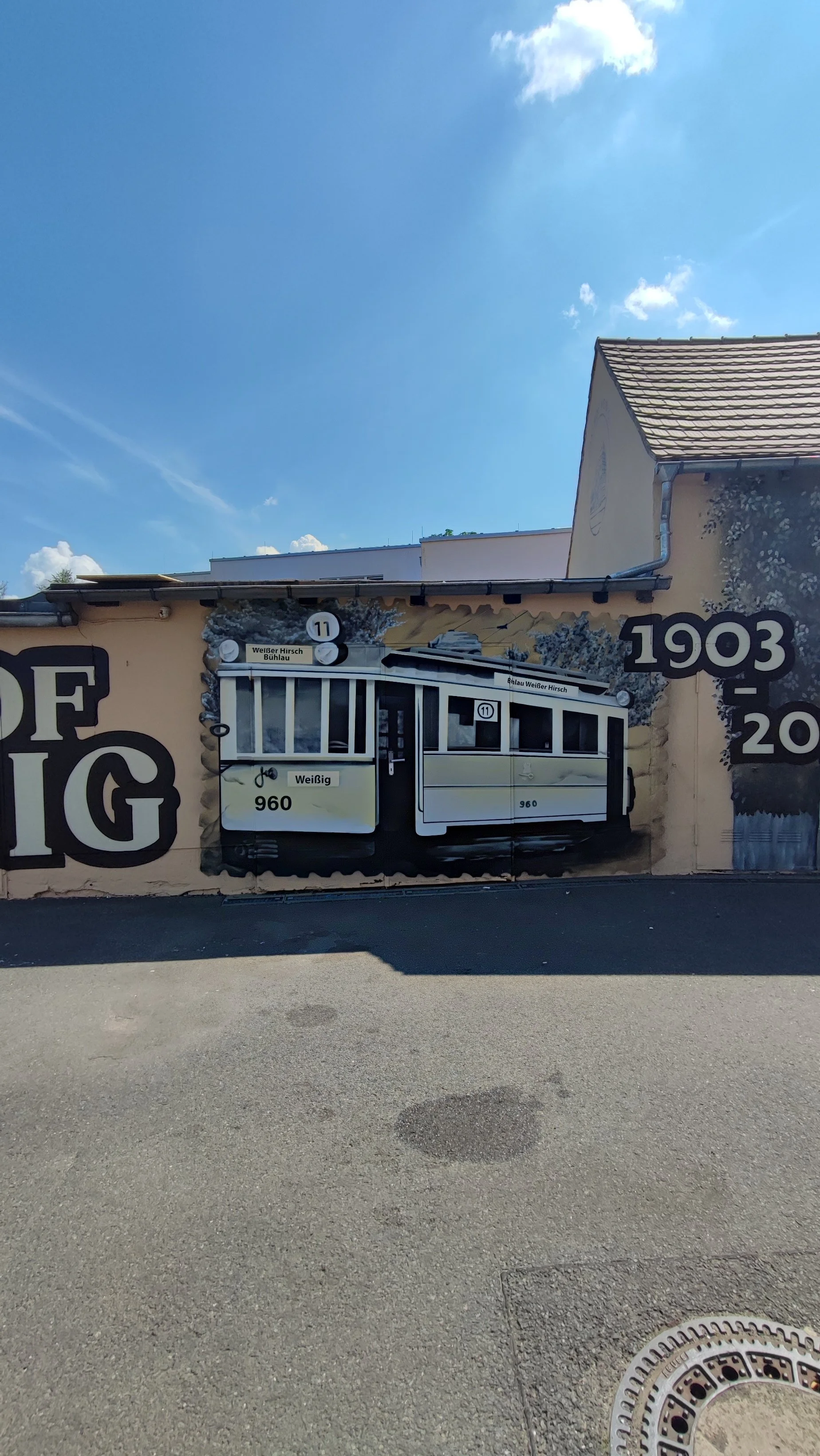Mural auf einer Wand, das einen Straßenbahnwagen darstellt, umgeben von Zahlen und Texten. Der Himmel ist blau mit wenigen Wolken.