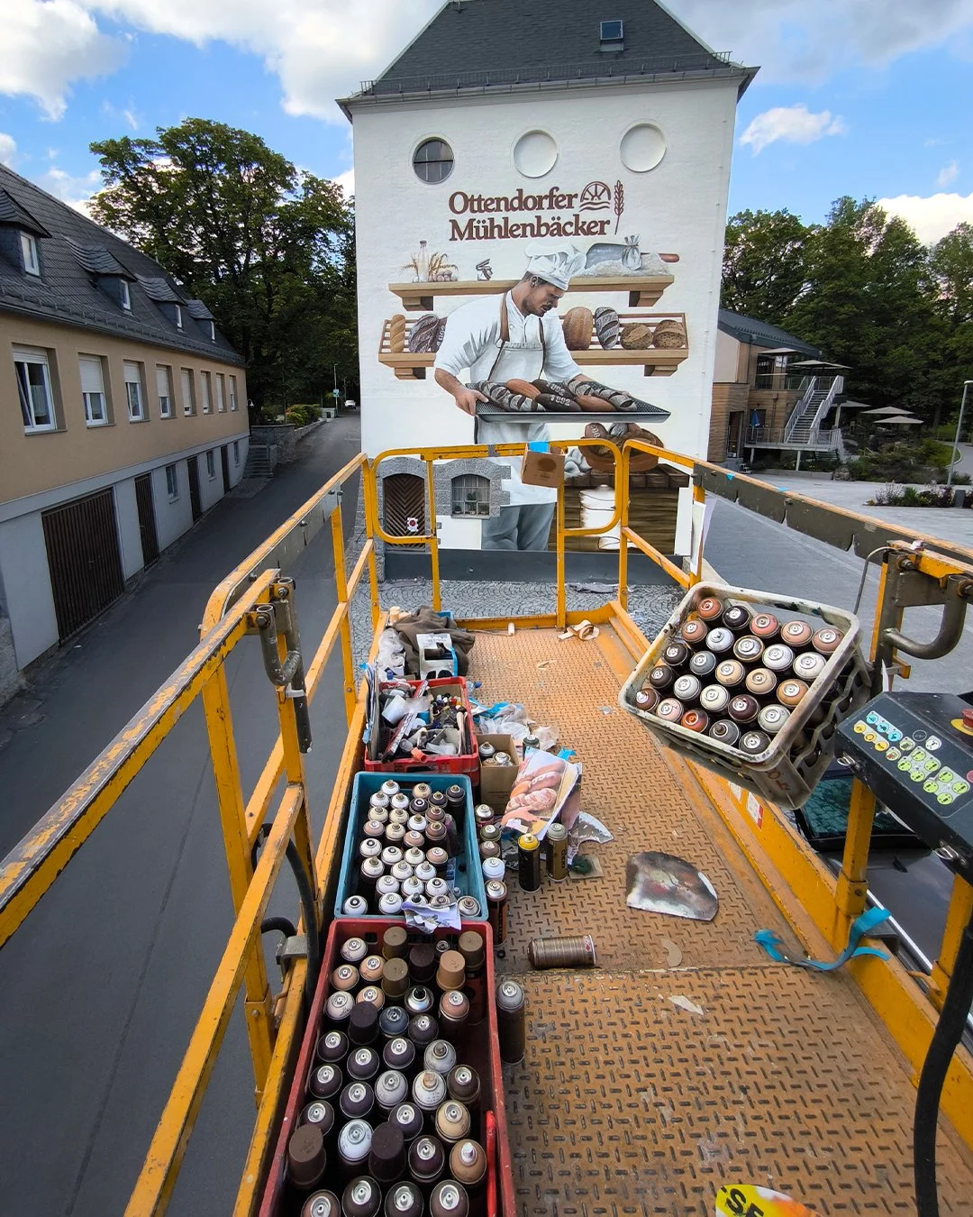 Auf einer gerüstartigen Plattform befinden sich Spraydosen, Werkzeuge und Farben, neben einem grauen Betonboden. Im Hintergrund ist ein großer Wandbild mit einem Bäcker bei der Arbeit in einer Bäckerei zu sehen, auf dem die Beschriftung "Ottendorfer 